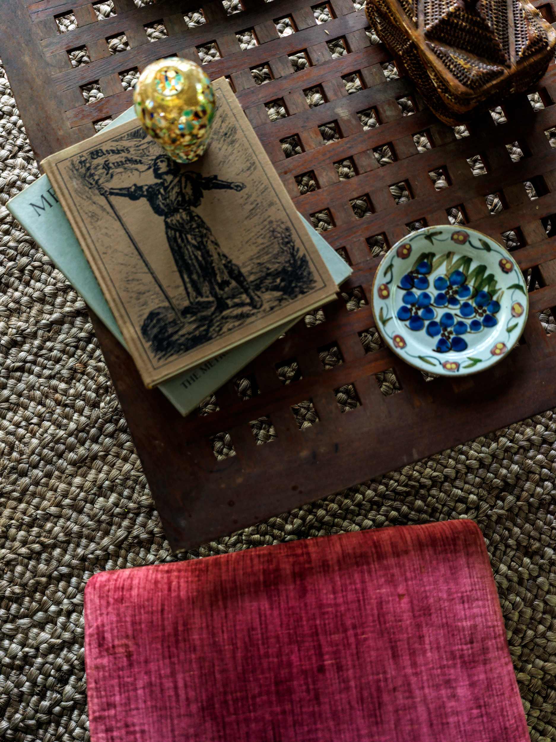 Close-up of a rustic wooden coffee table displaying stacked books, a decorative golden egg, and a small blue and white painted dish, with a plush red ottoman in the foreground and a woven rug beneath.