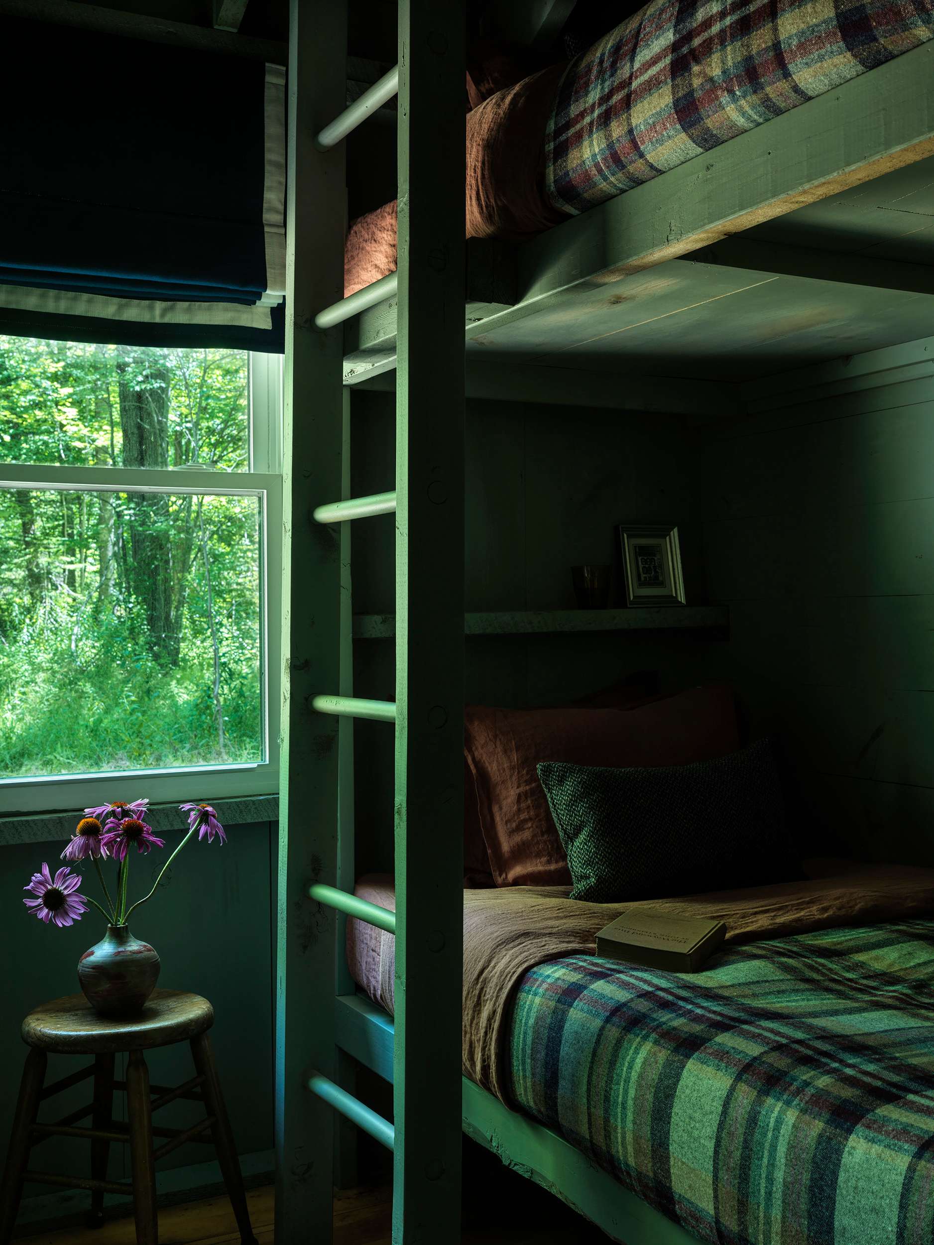 A cozy bunk bed room features dark green paneled walls, plaid bedding, and a large window overlooking lush green trees. A small wooden stool with a vase of purple flowers adds a touch of nature.