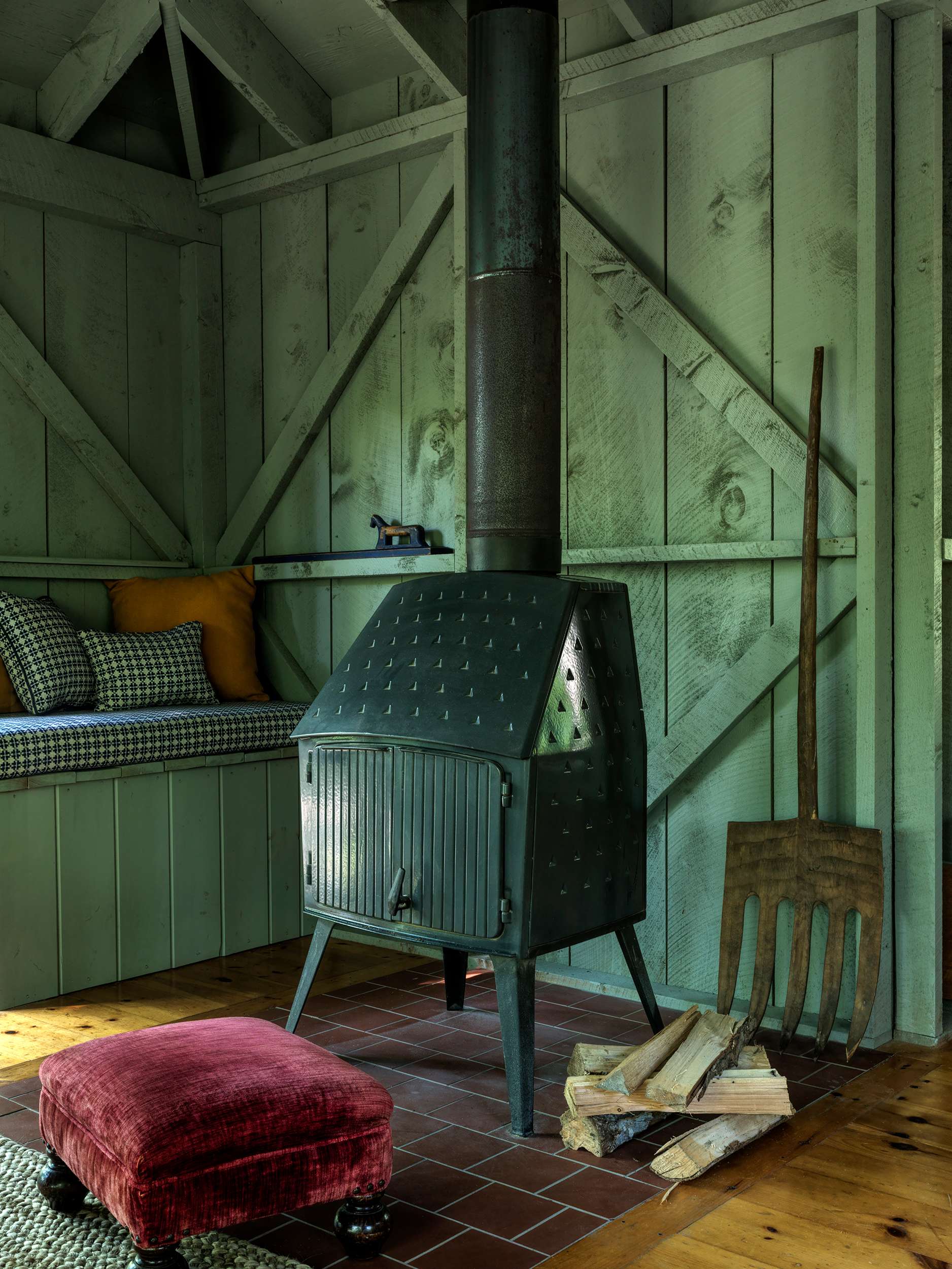 A cozy corner featuring a black wood-burning stove on terracotta tiles, flanked by light green wooden walls and a built-in bench with patterned cushions. A large wooden pitchfork leans against the wall.