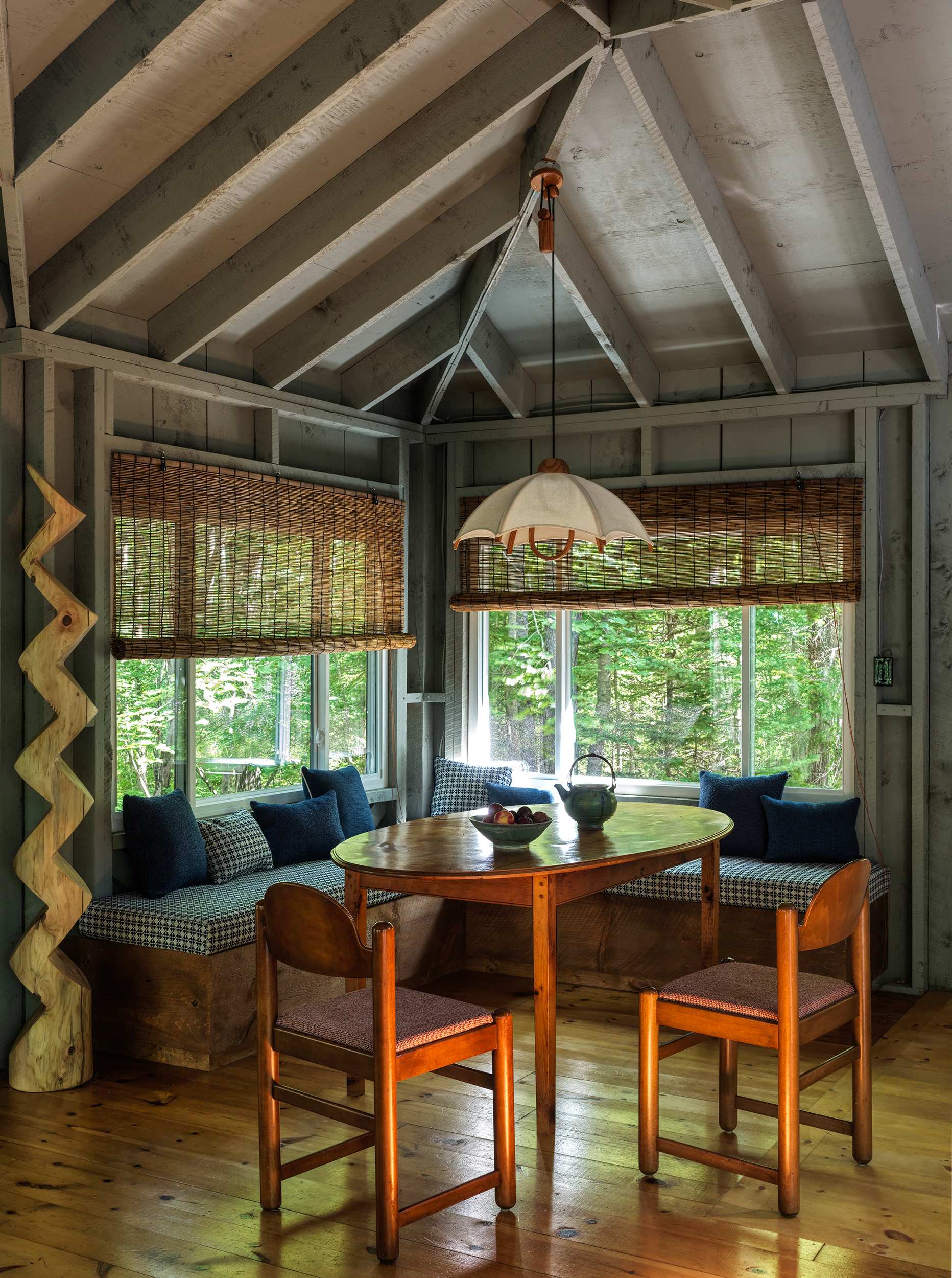 A bright dining area featuring a round wooden table surrounded by built-in bench seating with patterned cushions and two wooden chairs. Large windows with bamboo blinds offer views of the surrounding forest.