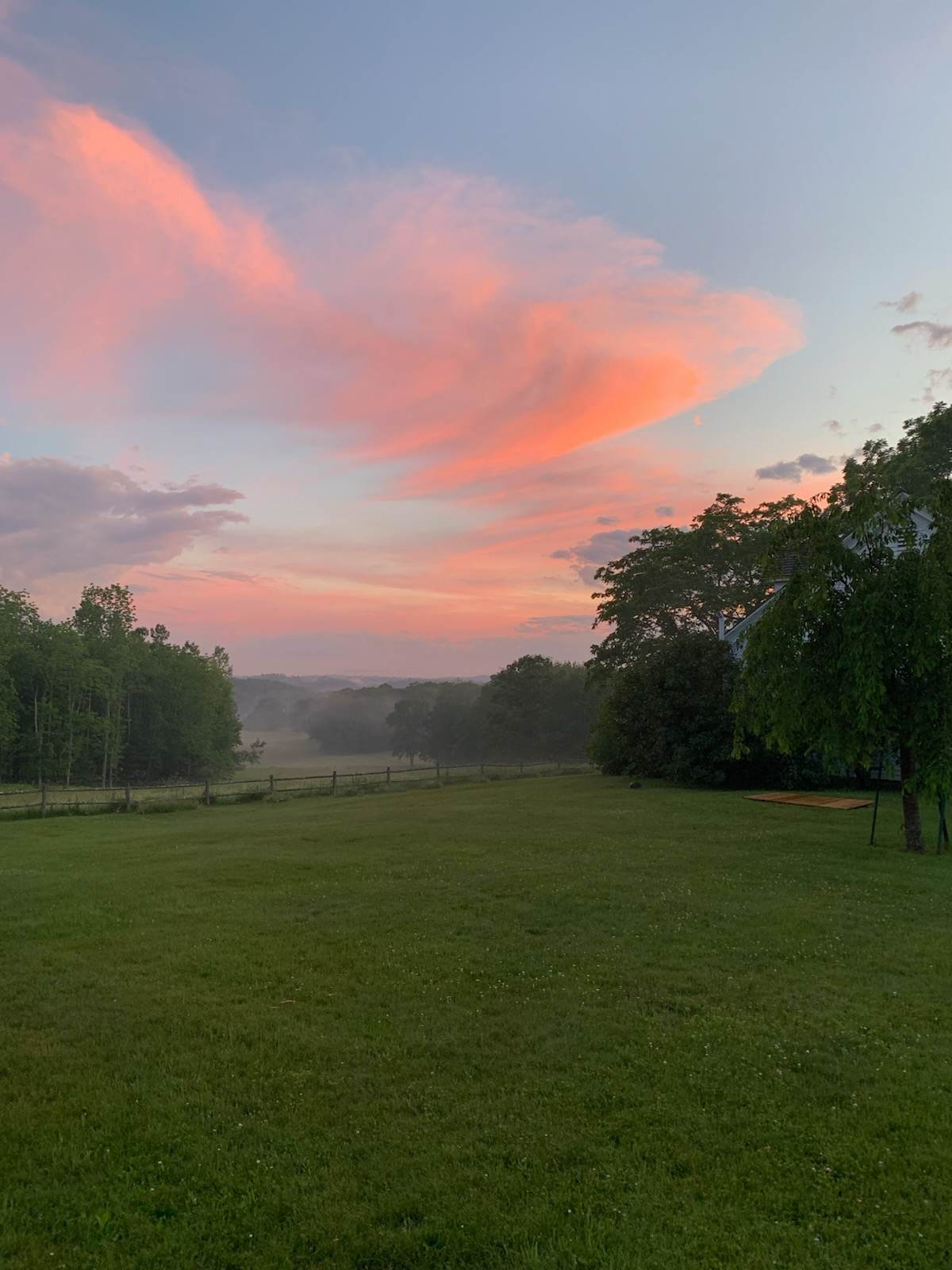 A sprawling green lawn extends towards a distant tree line and a misty valley, under a dramatic sunset sky painted with soft pink and orange clouds, creating a peaceful rural landscape.