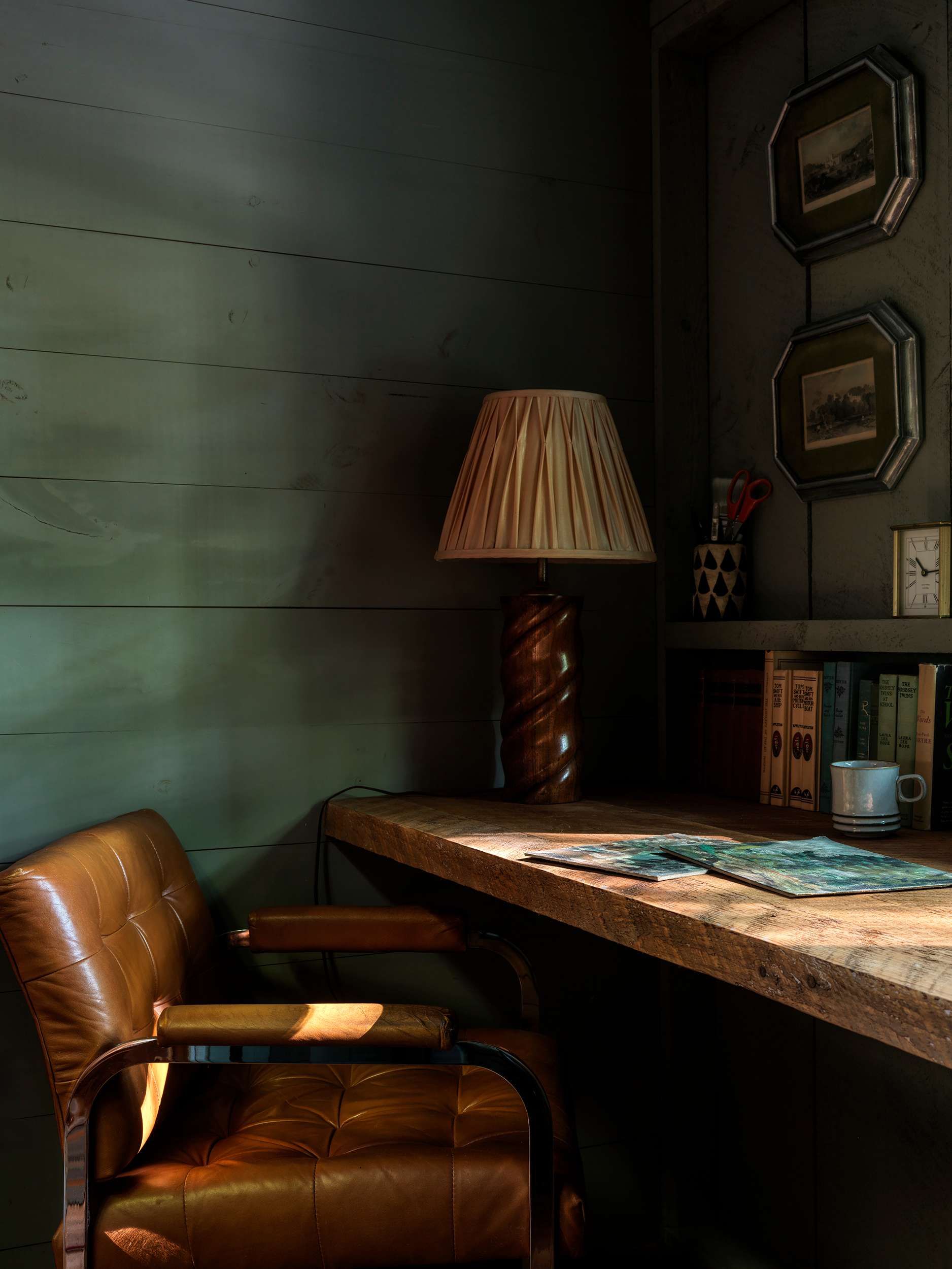 A rustic office nook with dark green paneled walls features a large wooden desk and a comfortable brown leather armchair. A table lamp with a pleated shade and framed artwork create a cozy workspace.