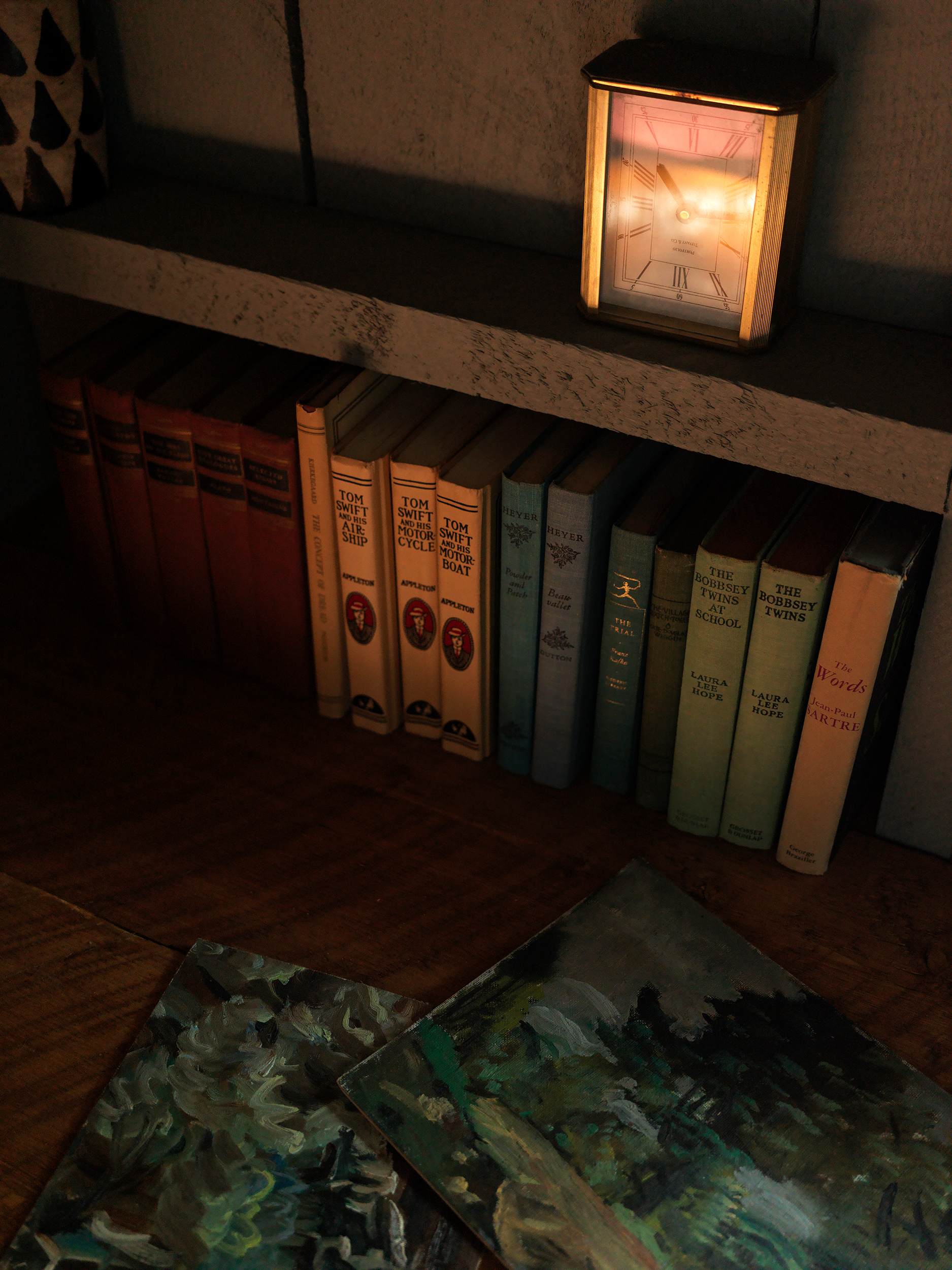 A close-up of a rustic wooden desk reveals vintage books neatly arranged on a shelf above, alongside a classic alarm clock and two small landscape paintings. The scene is dimly lit, highlighting the textures.
