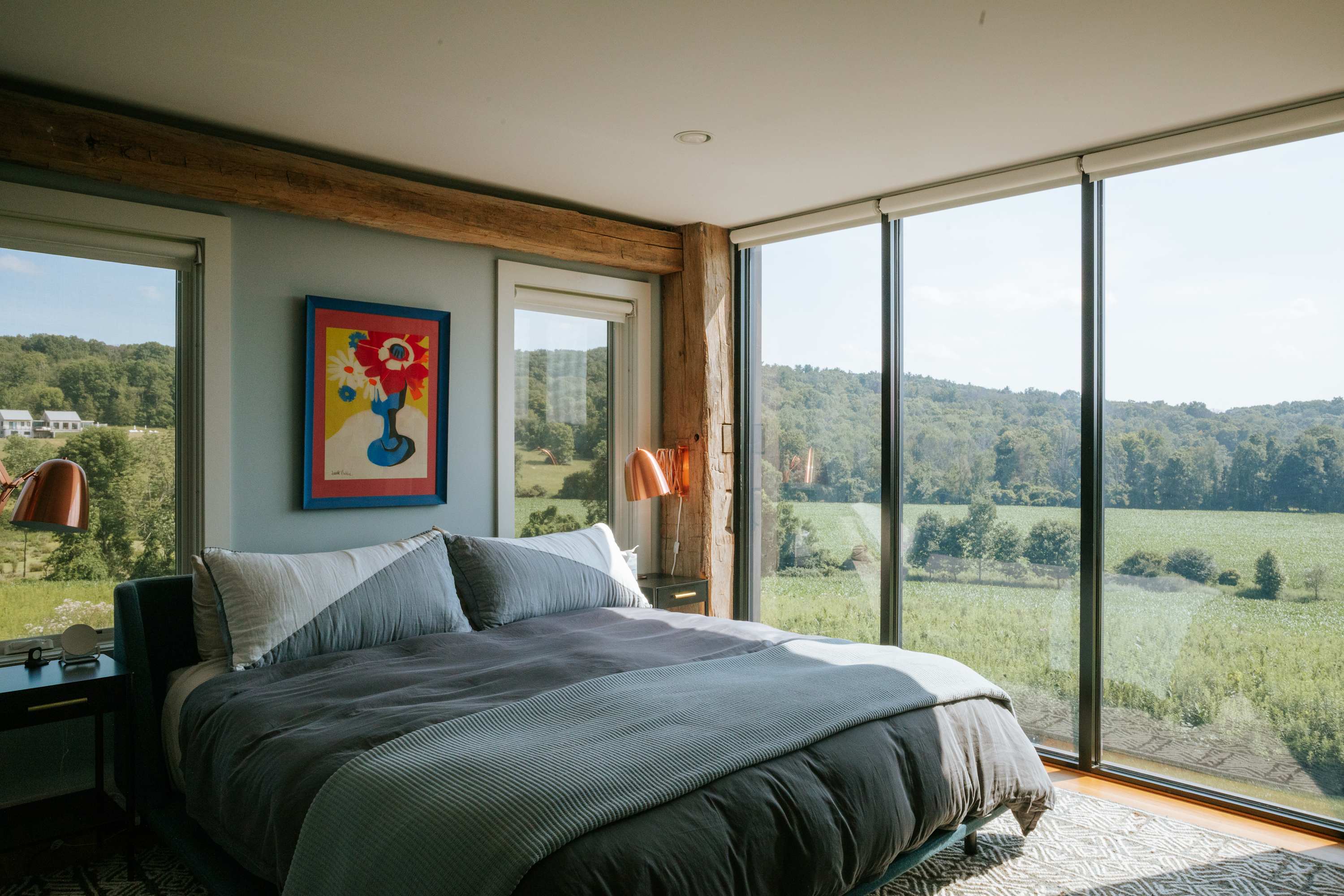 Sunlit bedroom with a large bed and gray linens, featuring a striking floor-to-ceiling window that provides expansive views of the surrounding green landscape. Exposed wooden beams add rustic charm, and a colorful artwork brightens the wall above the bed.