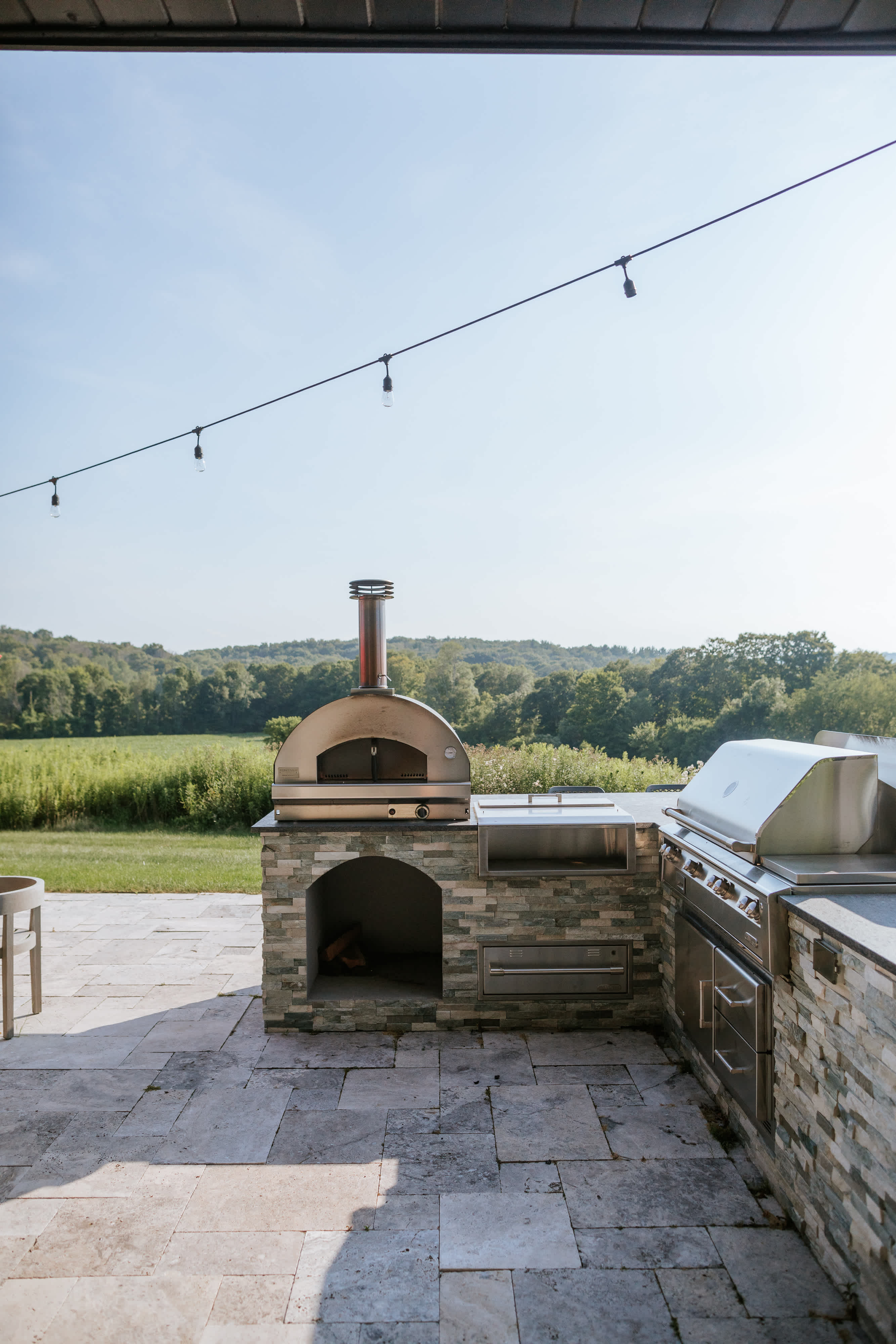 Outdoor kitchen area with a stone-clad base, featuring a stainless steel pizza oven with a tall chimney and a large built-in grill, set on a stone-tiled patio with string lights overhead and a green field in the background.