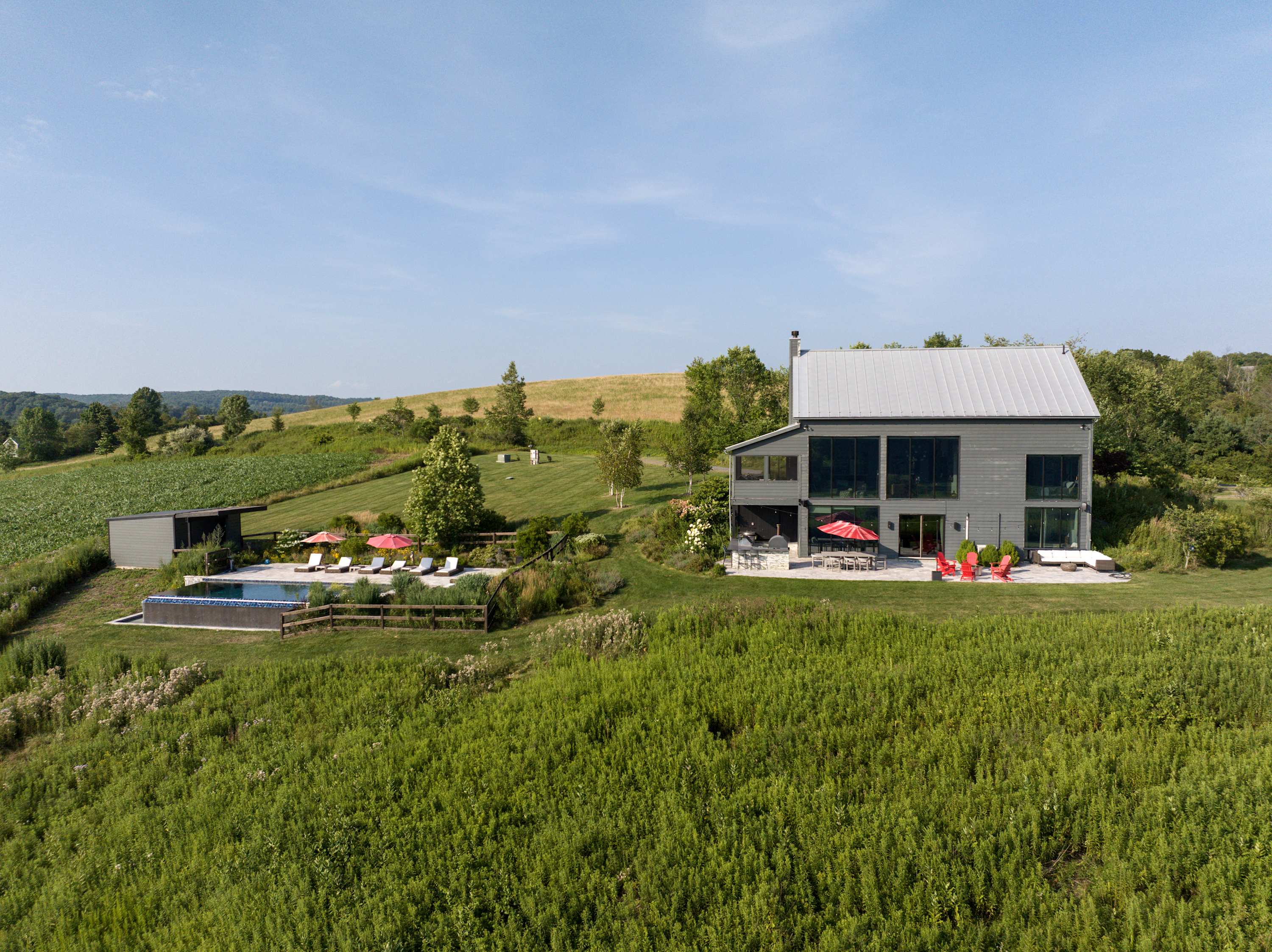 Aerial view of a modern gray barn-style house with a metal roof, large windows, and an adjacent infinity-edge pool area with lounge chairs and red umbrellas, all set within a vast green, hilly landscape.