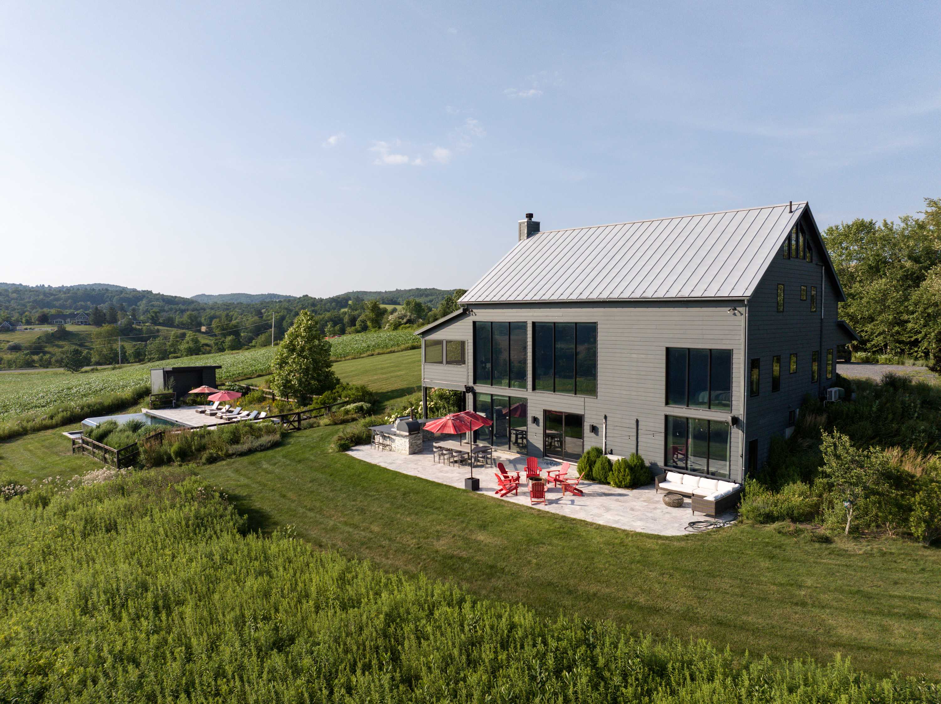 An aerial view showcases a modern gray barn-style house with a metal roof, surrounded by lush green fields and distant hills under a clear sky. A large patio with red Adirondack chairs, an outdoor kitchen, and a swimming pool are visible next to the house.