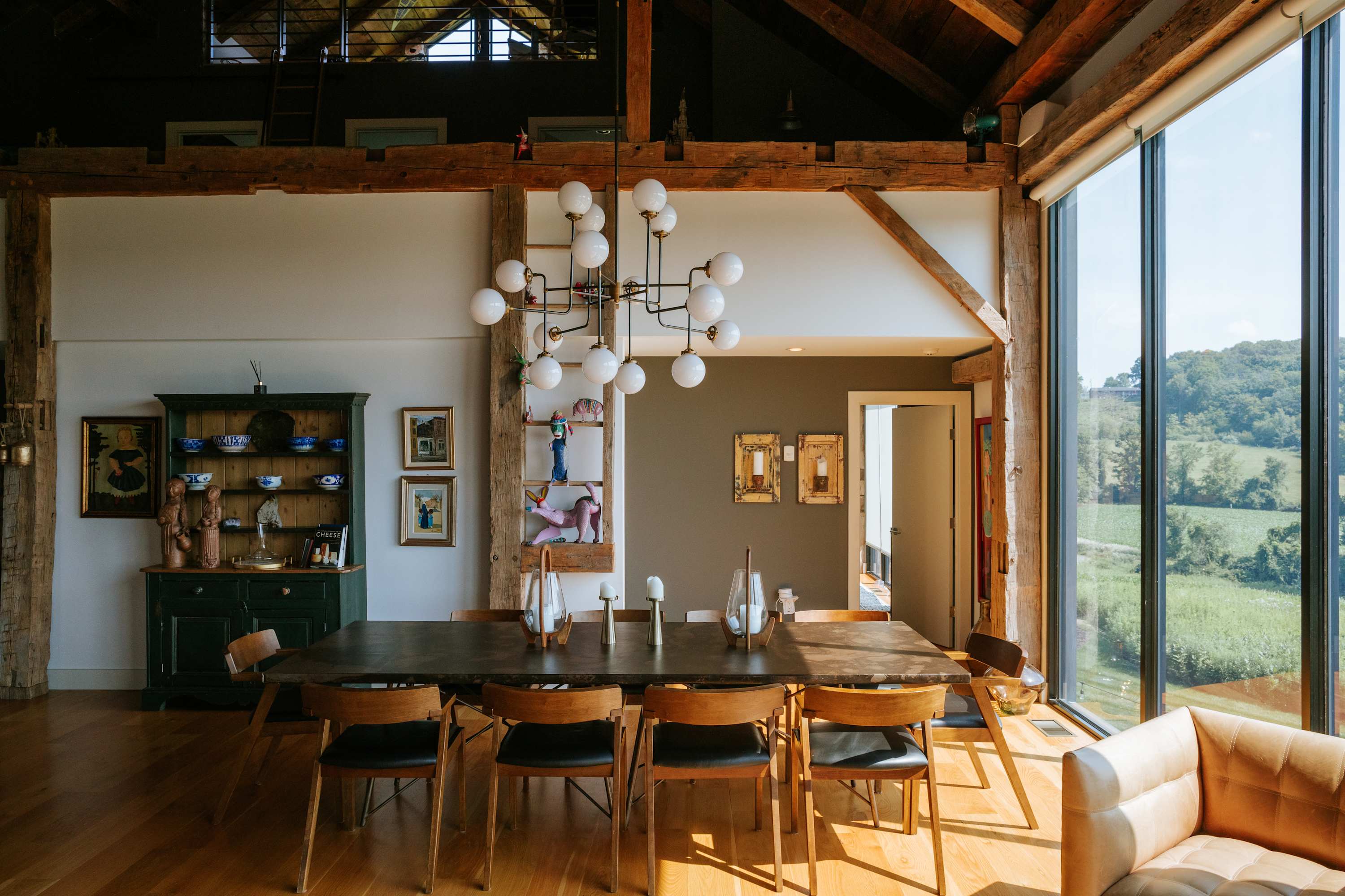 Spacious dining area with a long dark wood table and mid-century modern chairs, illuminated by a multi-globe chandelier and natural light from large windows. Rustic exposed timber beams frame the high ceiling, complementing the green hutch and various wall art.
