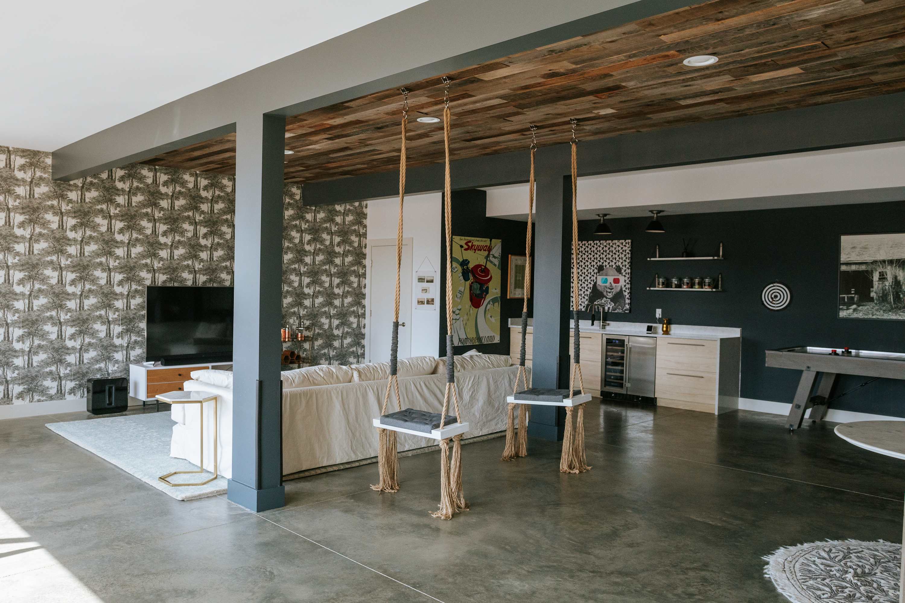 An expansive open-concept living and game room features a white sofa, a TV against a patterned accent wall, and two indoor swings. The space transitions into a wet bar area with a pool table, all under a reclaimed wood ceiling and dark gray structural beams.