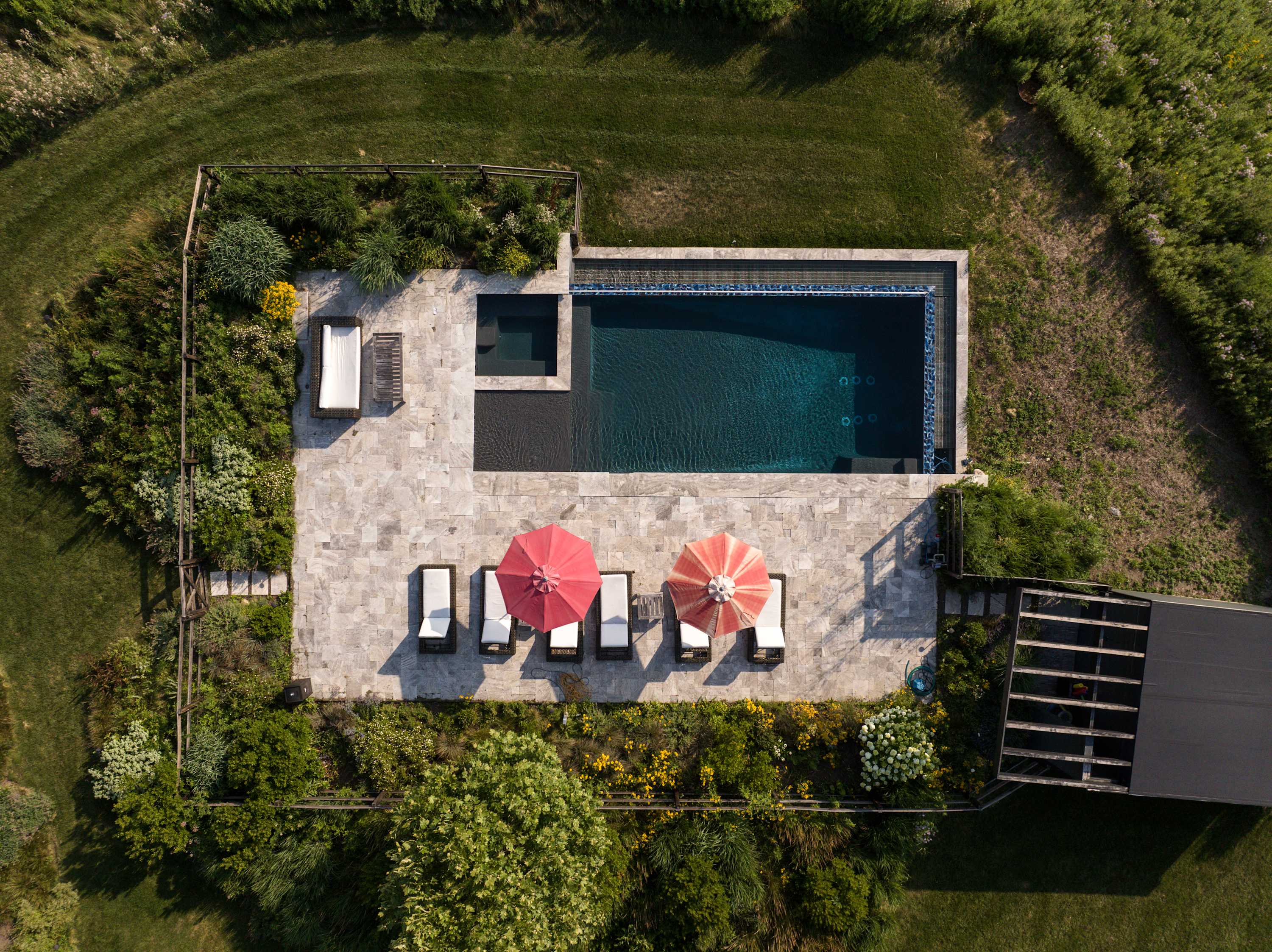 Overhead aerial view of the rectangular infinity-edge swimming pool with dark blue tiles, surrounded by a light-colored stone patio, lounge chairs, and red umbrellas, with a small pool house nearby.