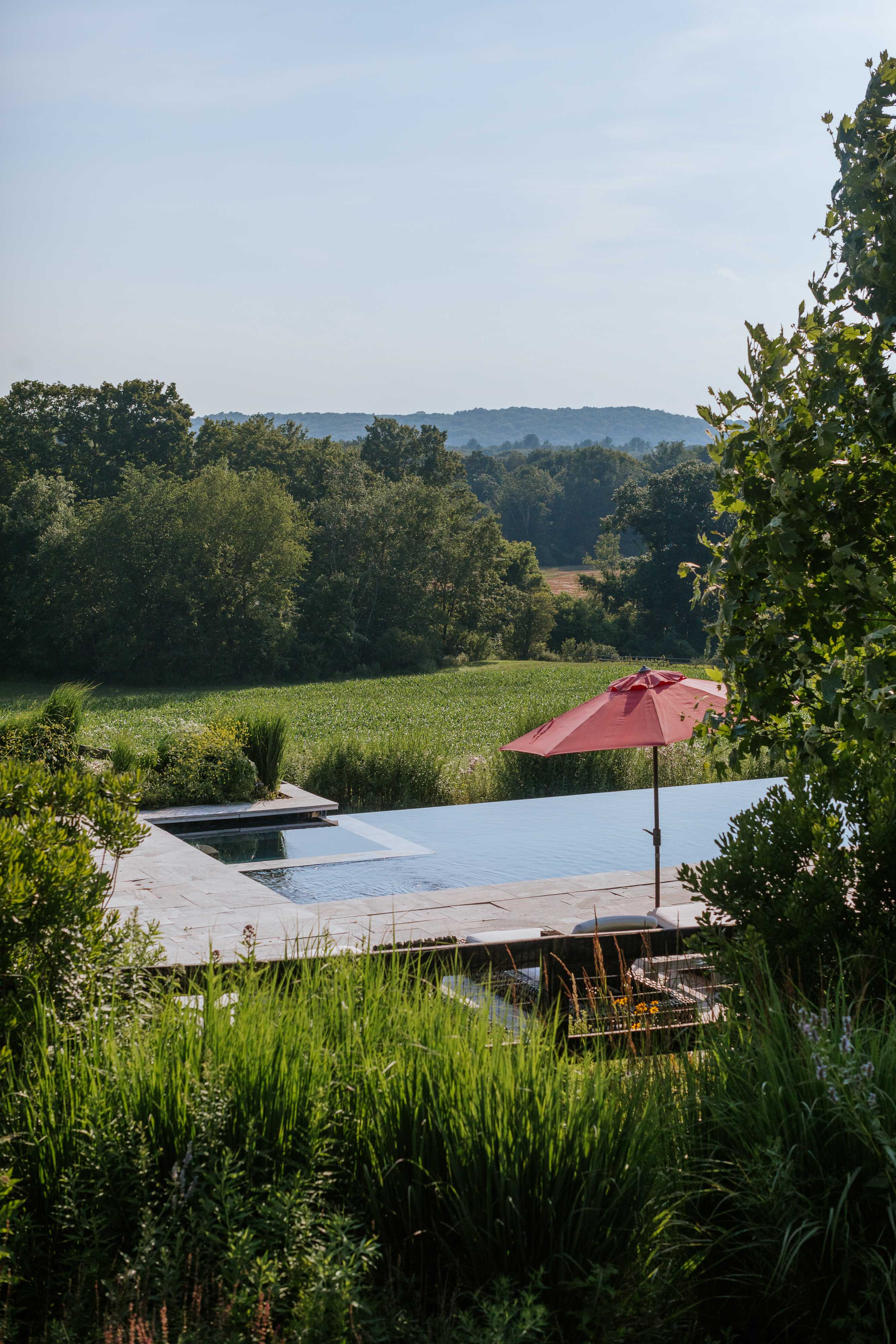 Expansive infinity pool with a red umbrella on the stone deck, offering panoramic views of a sprawling green field and rolling hills, with lush foreground vegetation.