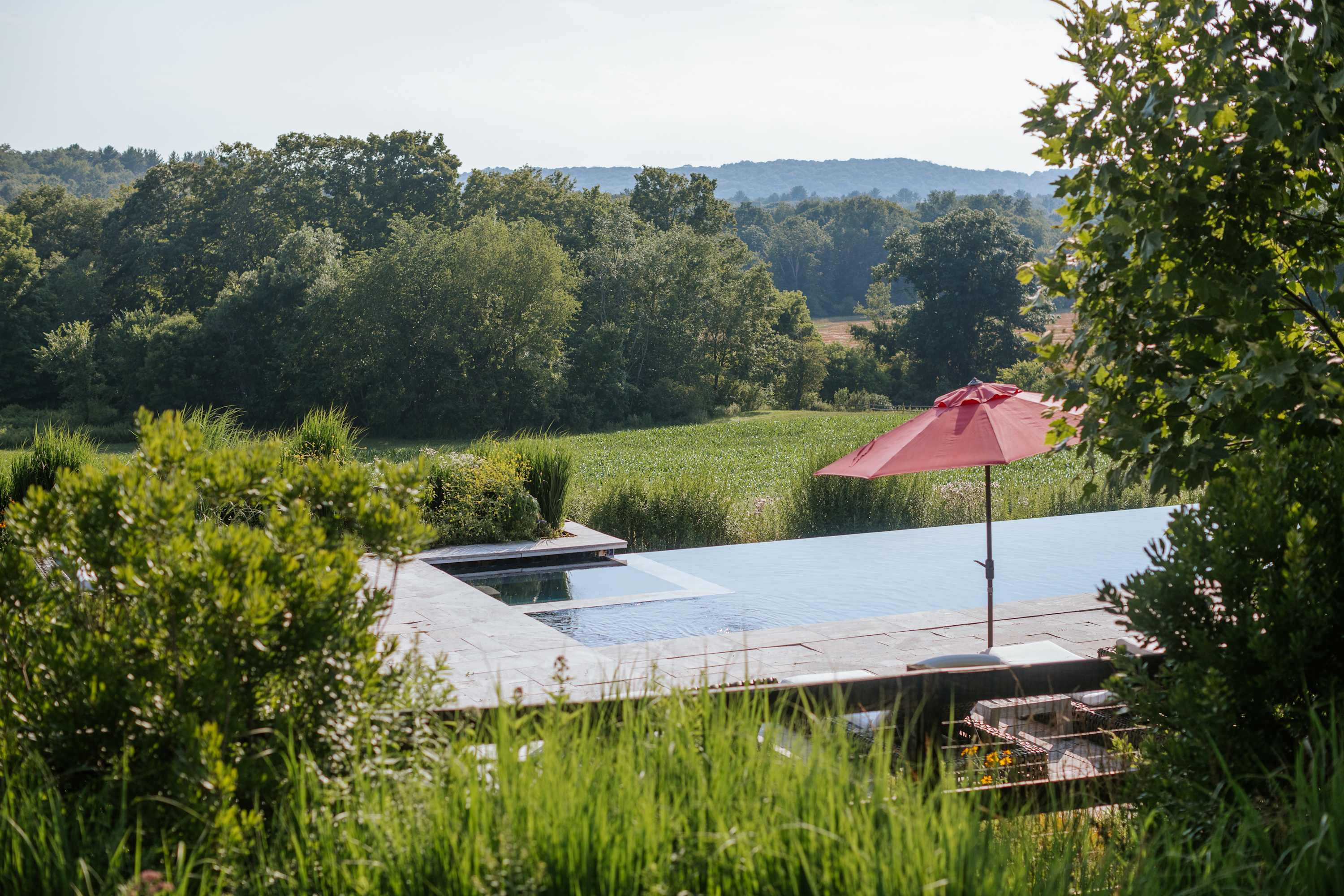 Serene infinity pool with a red umbrella on the deck, overlooking a vast green field and distant tree-covered hills under a clear sky, framed by dense foliage in the foreground.