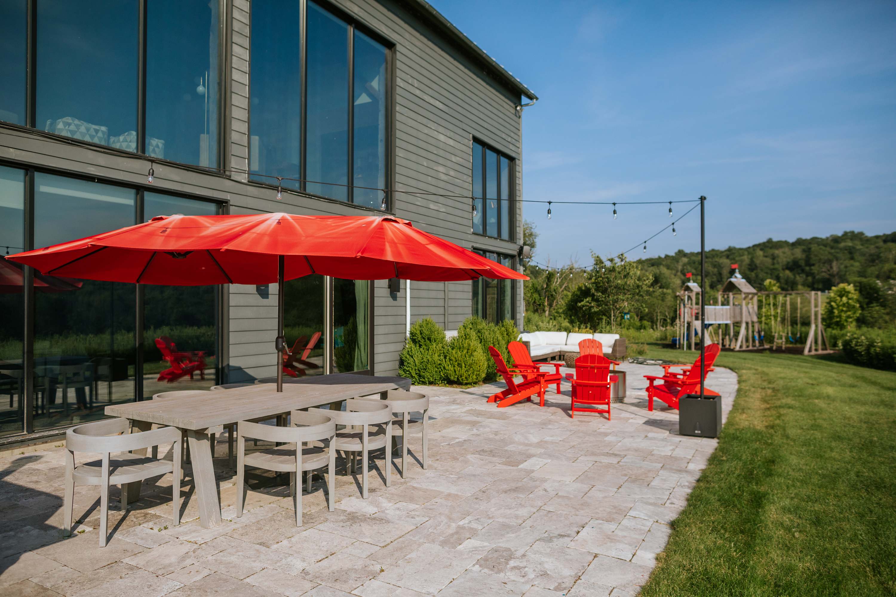 Outdoor dining area on a light-colored stone patio, featuring a long wooden dining table with gray chairs, a large red umbrella, and string lights overhead, with the modern house and green landscape in the background.