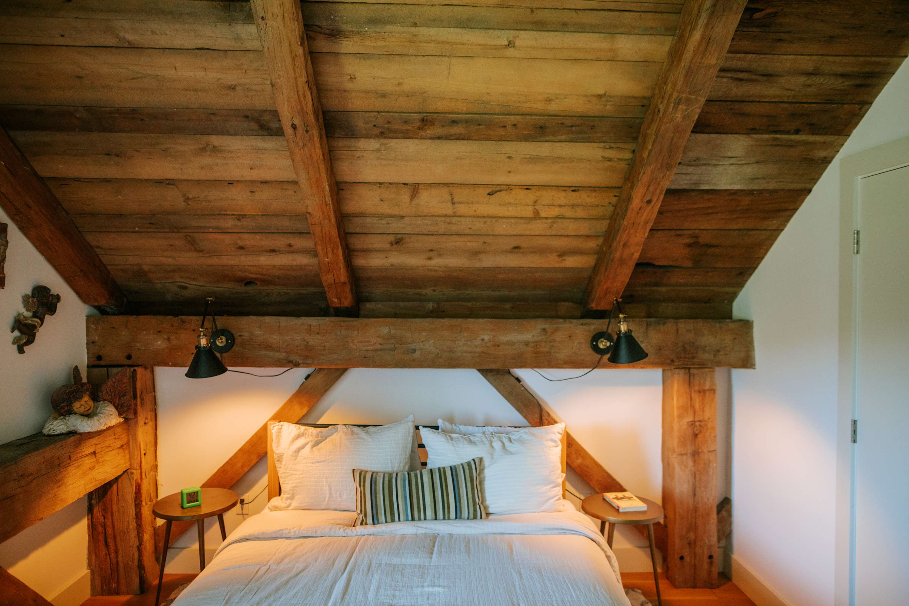 A rustic bedroom features a queen-sized bed with white bedding and a striped accent pillow, flanked by round wooden nightstands with small lamps. Exposed wooden beams form a striking headboard and ceiling, creating a warm, inviting atmosphere.