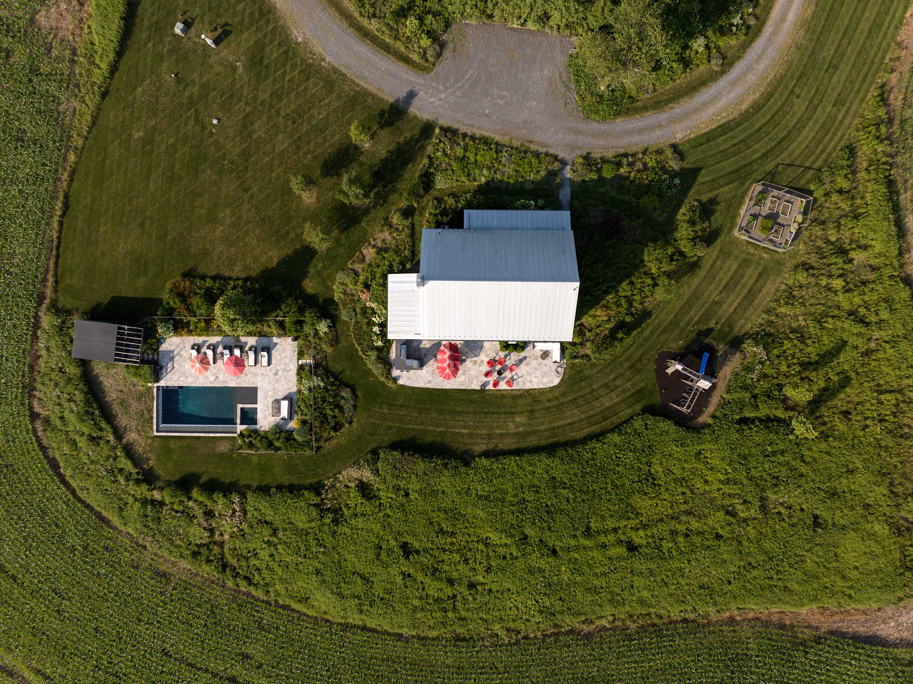 Overhead aerial view showcasing the gray barn-style house with its metal roof, a large stone patio with red umbrellas, an infinity pool, and a wooden playground, all nestled within lush green fields.