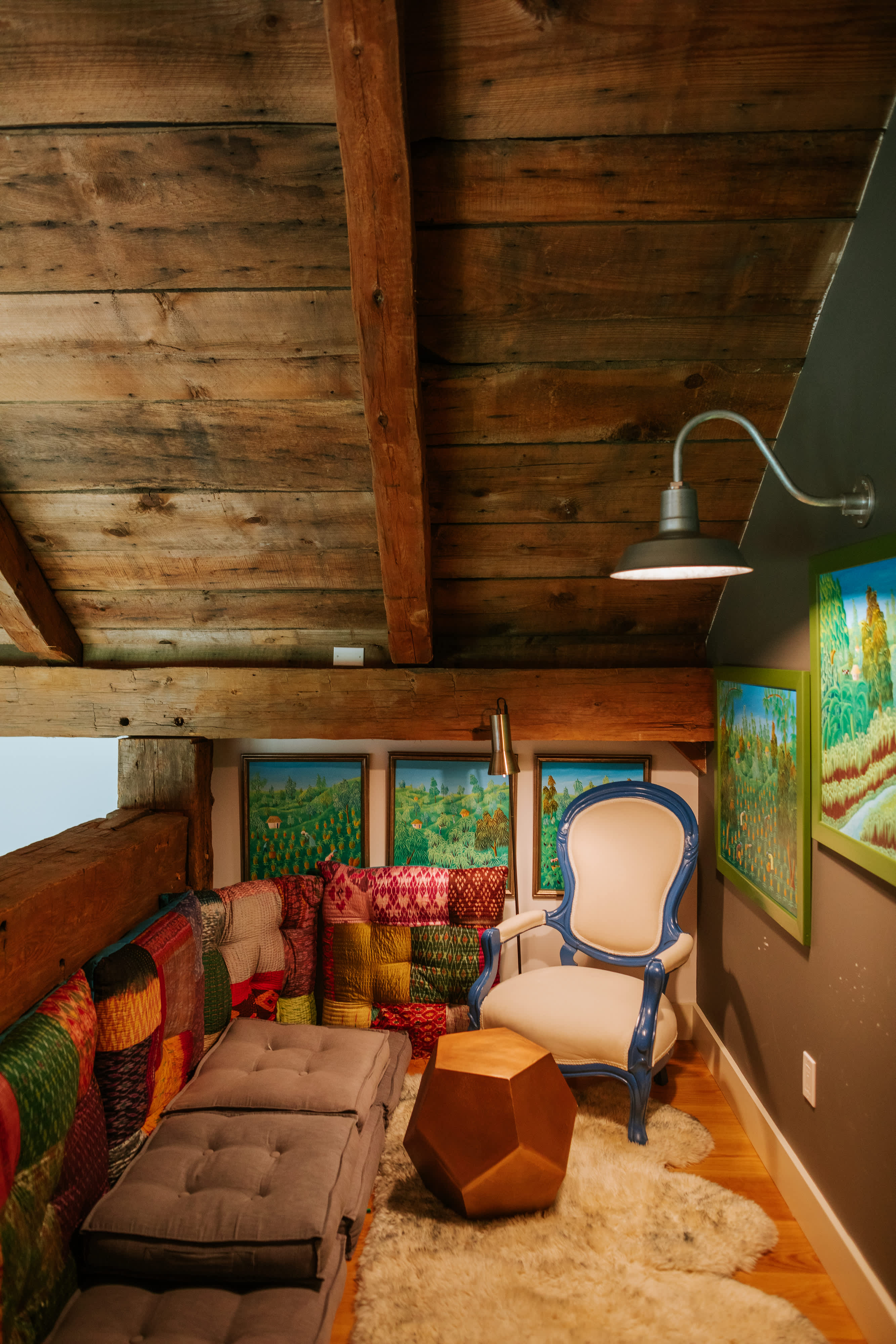 A cozy loft nook features a low sofa with colorful patterned cushions and floor pillows, a blue and white armchair, and a geometric wooden side table on a shaggy rug. Exposed wooden beams on the ceiling and walls add rustic charm, complemented by framed landscape paintings.