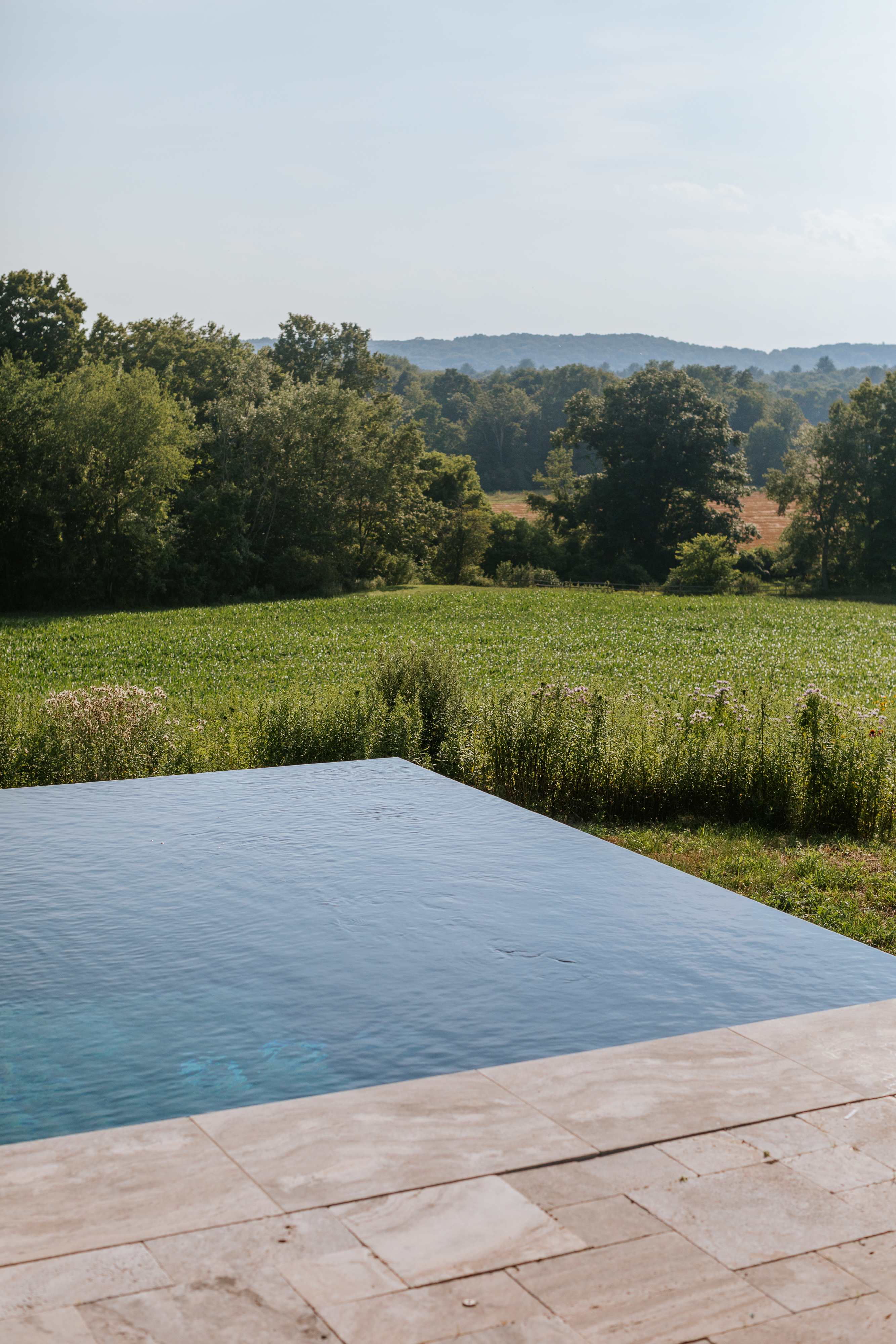 Close-up view of the tranquil infinity pool's edge, showcasing the clear blue water against a backdrop of a vibrant green field and distant tree-lined hills under a bright sky.