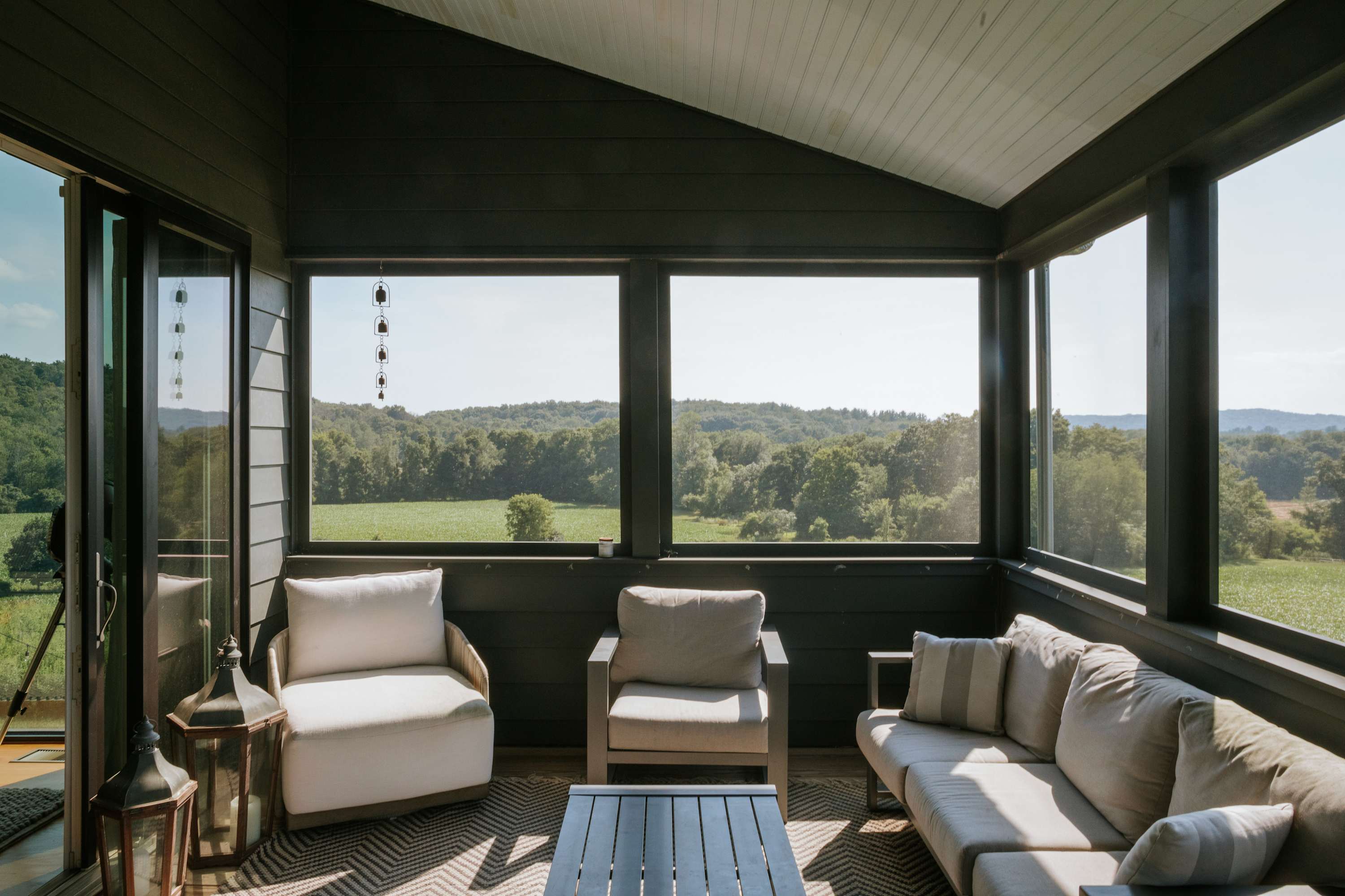 A cozy screened porch with dark gray walls and ceiling offers comfortable outdoor seating, including a sofa and armchairs with light cushions, providing a sheltered space to enjoy the expansive green landscape views.