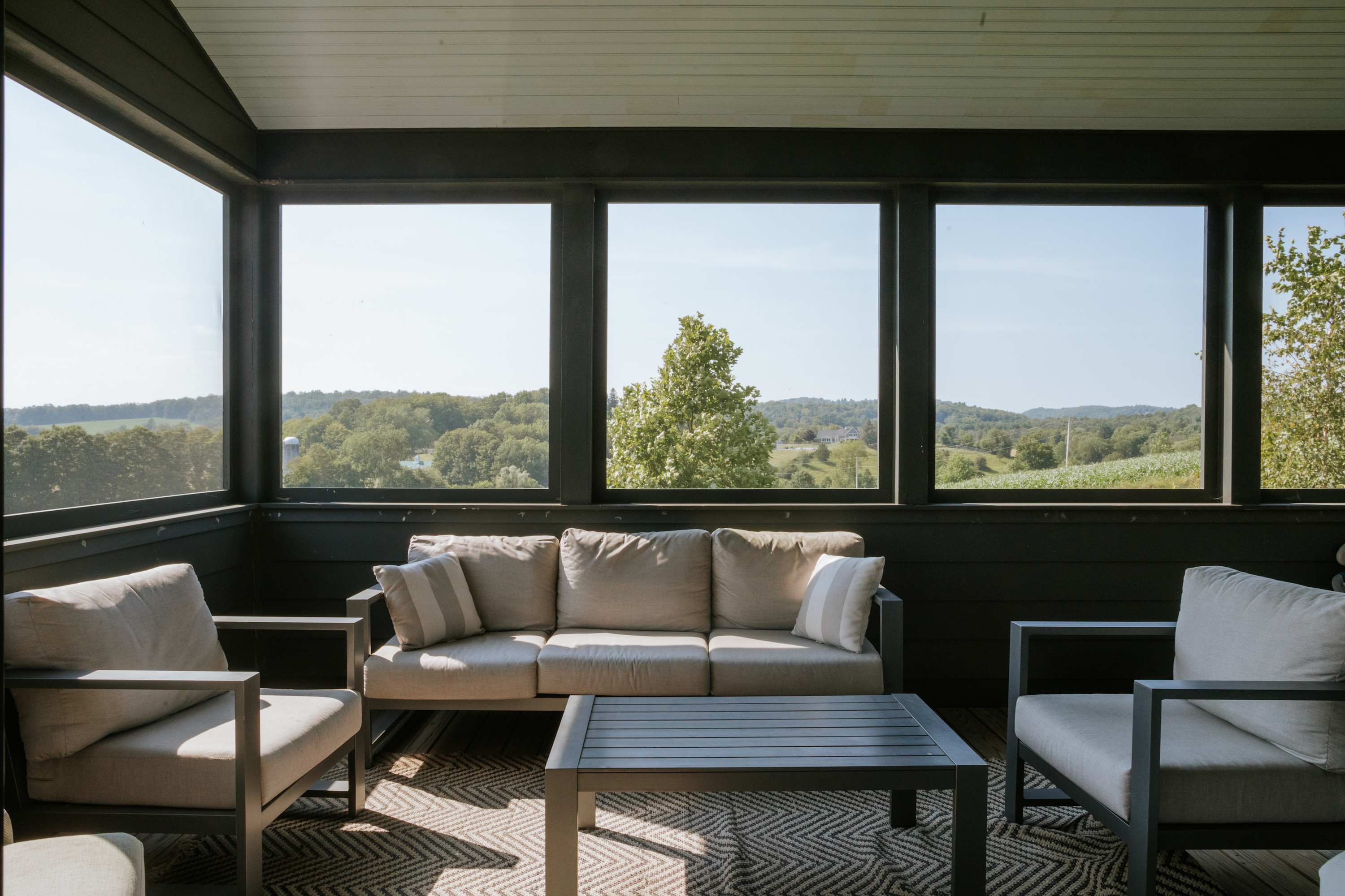 A screened-in porch features comfortable outdoor seating, including a sofa and armchairs with light gray cushions, arranged around a dark slatted coffee table. Large windows offer panoramic views of rolling green hills and distant trees.