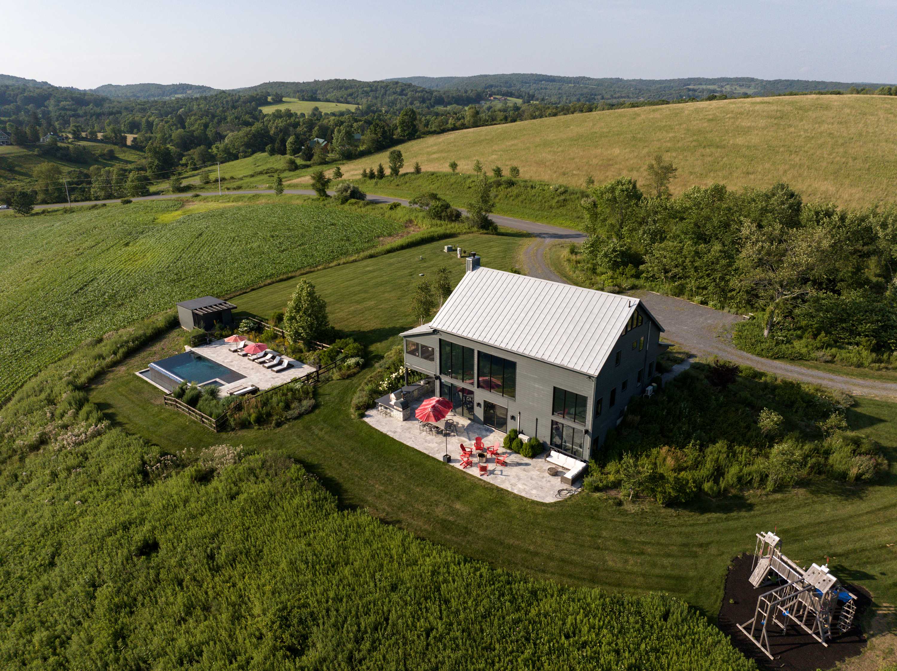 Wide aerial view of the modern gray house with a metal roof, featuring a large patio with red umbrellas, an infinity pool, a wooden playground, and a winding gravel driveway, all set against a backdrop of rolling green hills.