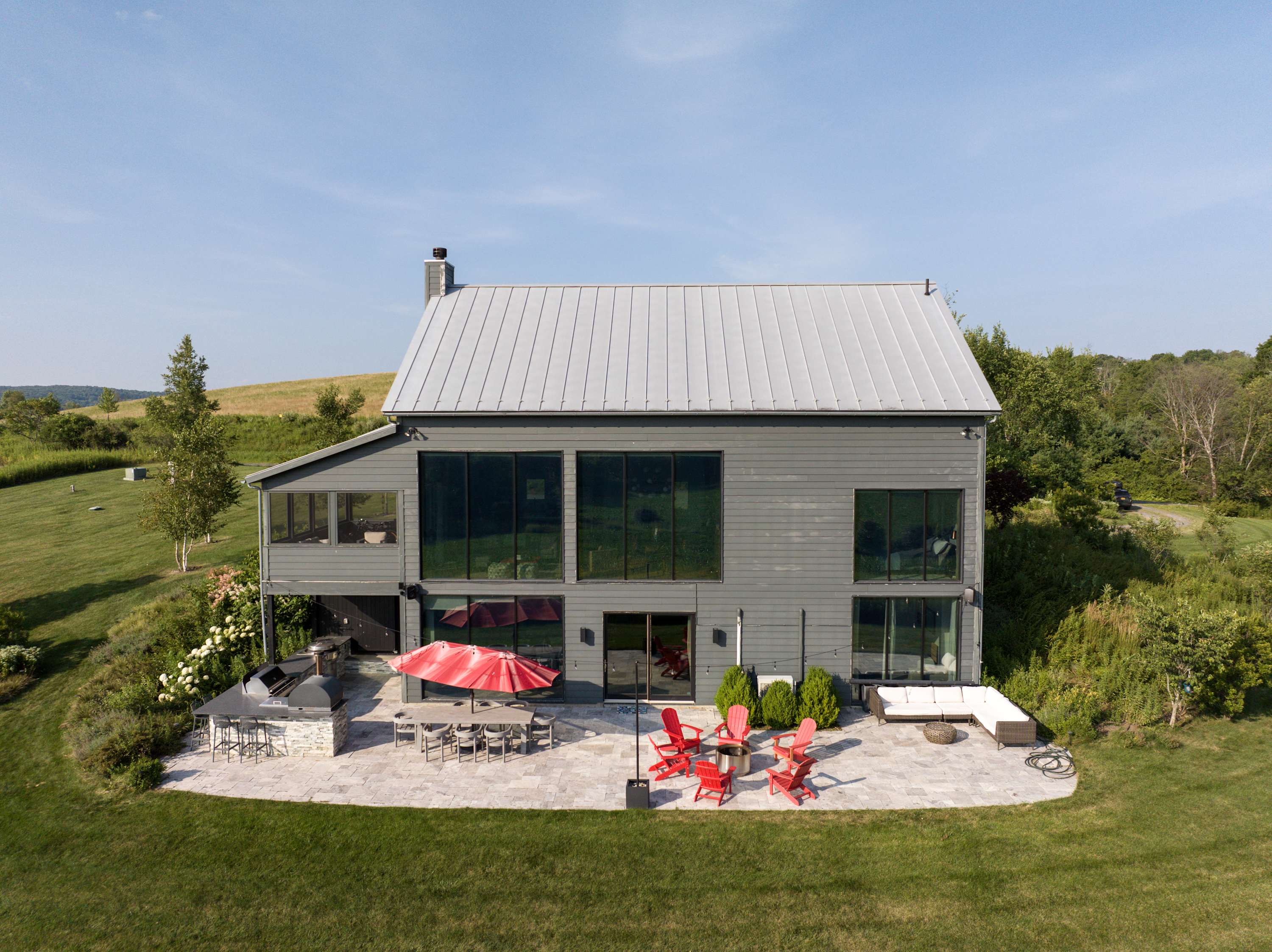 Rear exterior of the modern gray barn-style house with large windows, showcasing a spacious stone patio furnished with an outdoor dining table, red umbrella, BBQ grill, and a fire pit area with red Adirondack chairs.