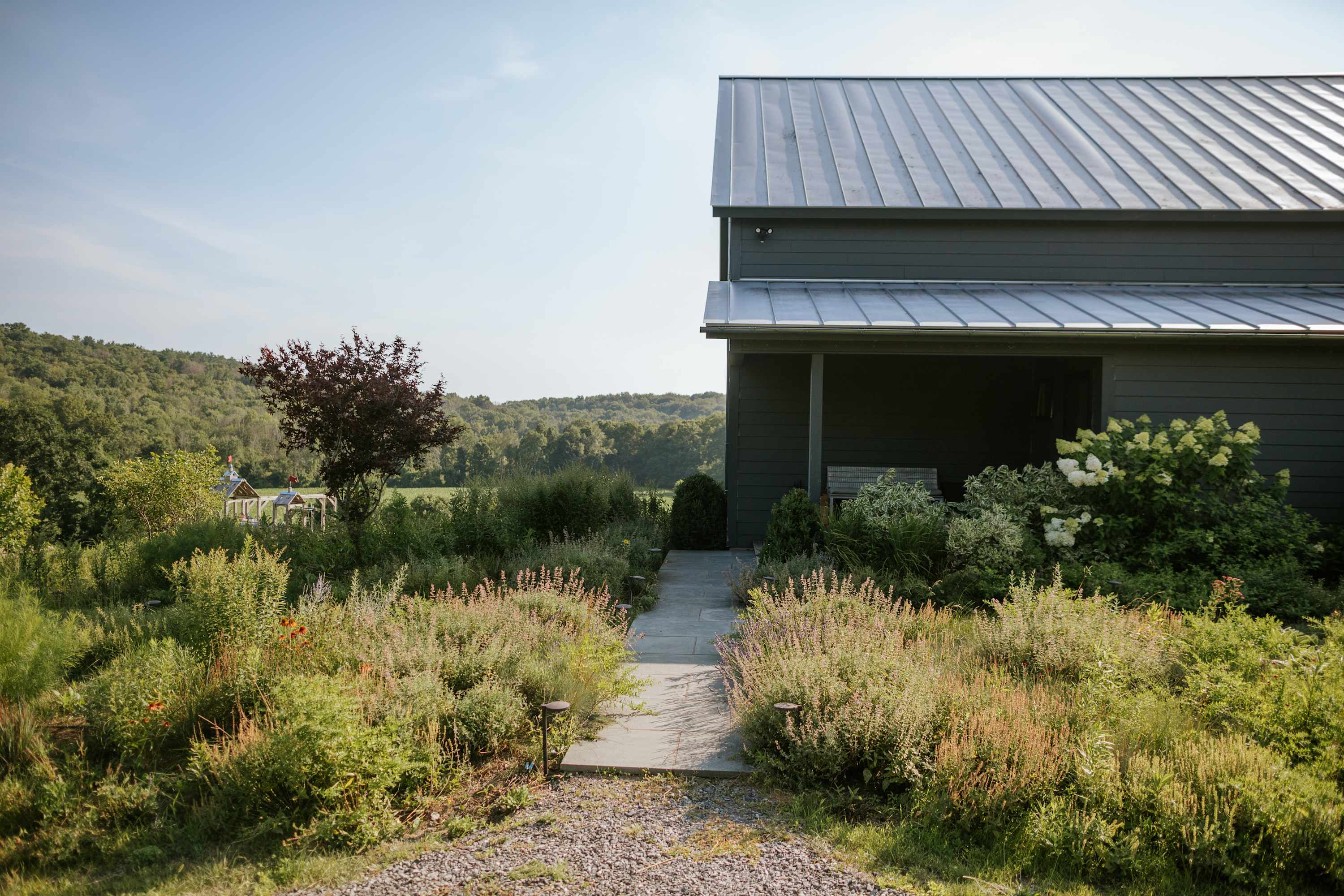 The side exterior of a modern gray barn-style house with a metal roof is framed by a stone pathway leading through a vibrant garden filled with various plants and shrubs, offering a glimpse of the distant green landscape.