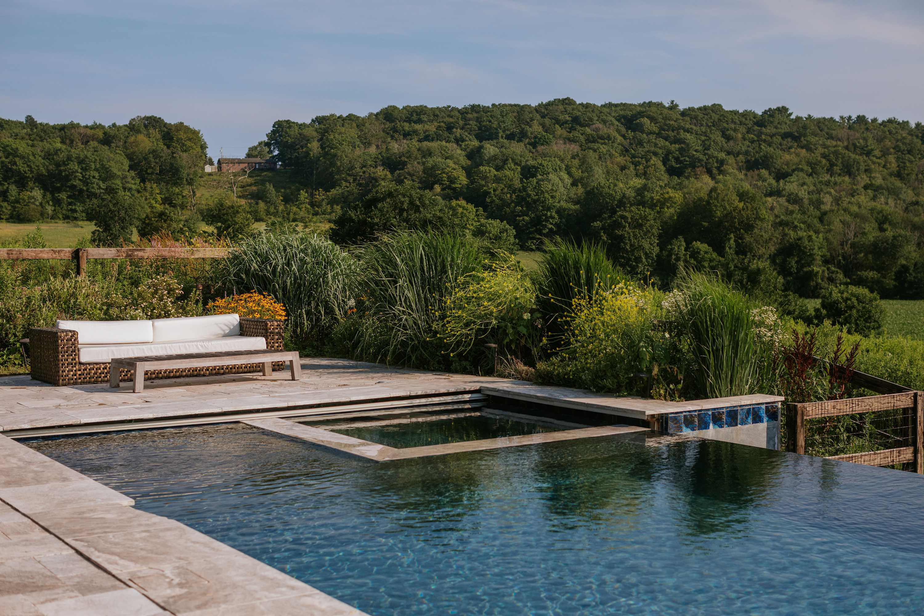 Outdoor pool area featuring an infinity pool, an integrated hot tub, and a wicker sofa with white cushions on a stone patio, surrounded by diverse plantings and a wooded hillside.