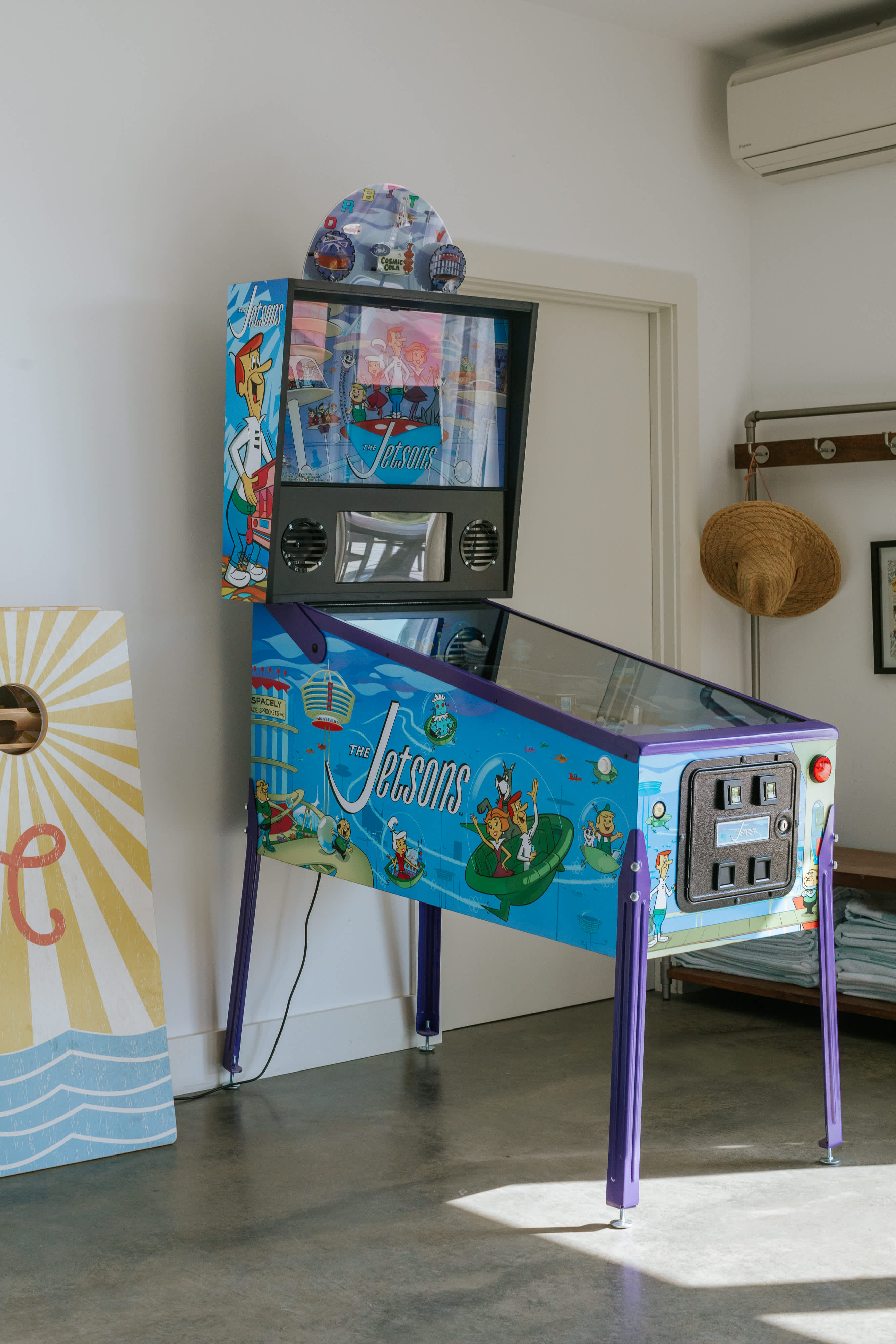 A vibrant "The Jetsons" themed pinball machine stands prominently in a bright room with white walls and a concrete floor. A cornhole board is partially visible to the left, indicating a dedicated game area.
