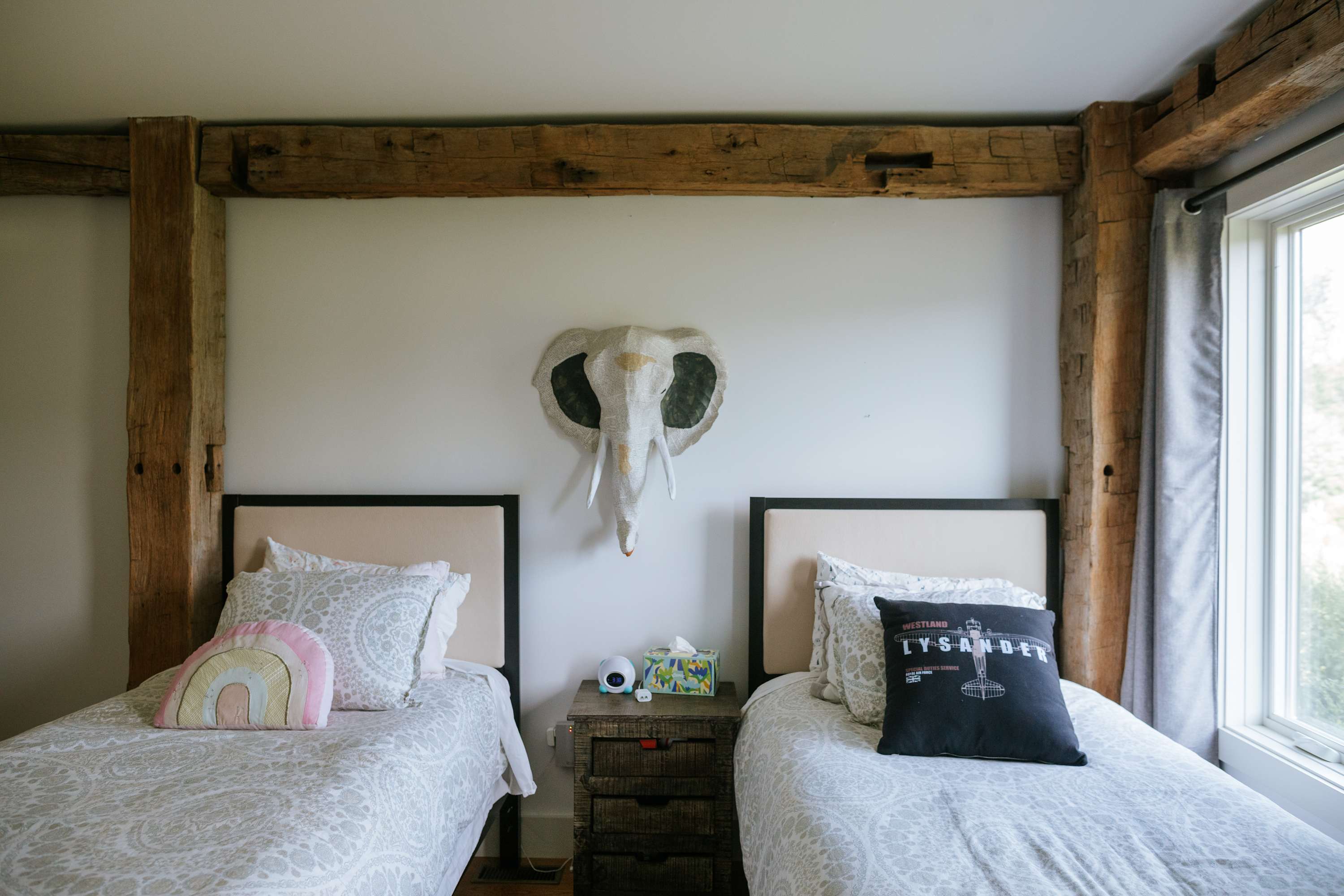 Detailed view of a children's bedroom featuring two twin beds with patterned bedding and a shared rustic nightstand. An artistic elephant head wall sculpture hangs above the beds, adding a playful touch to the room's decor.