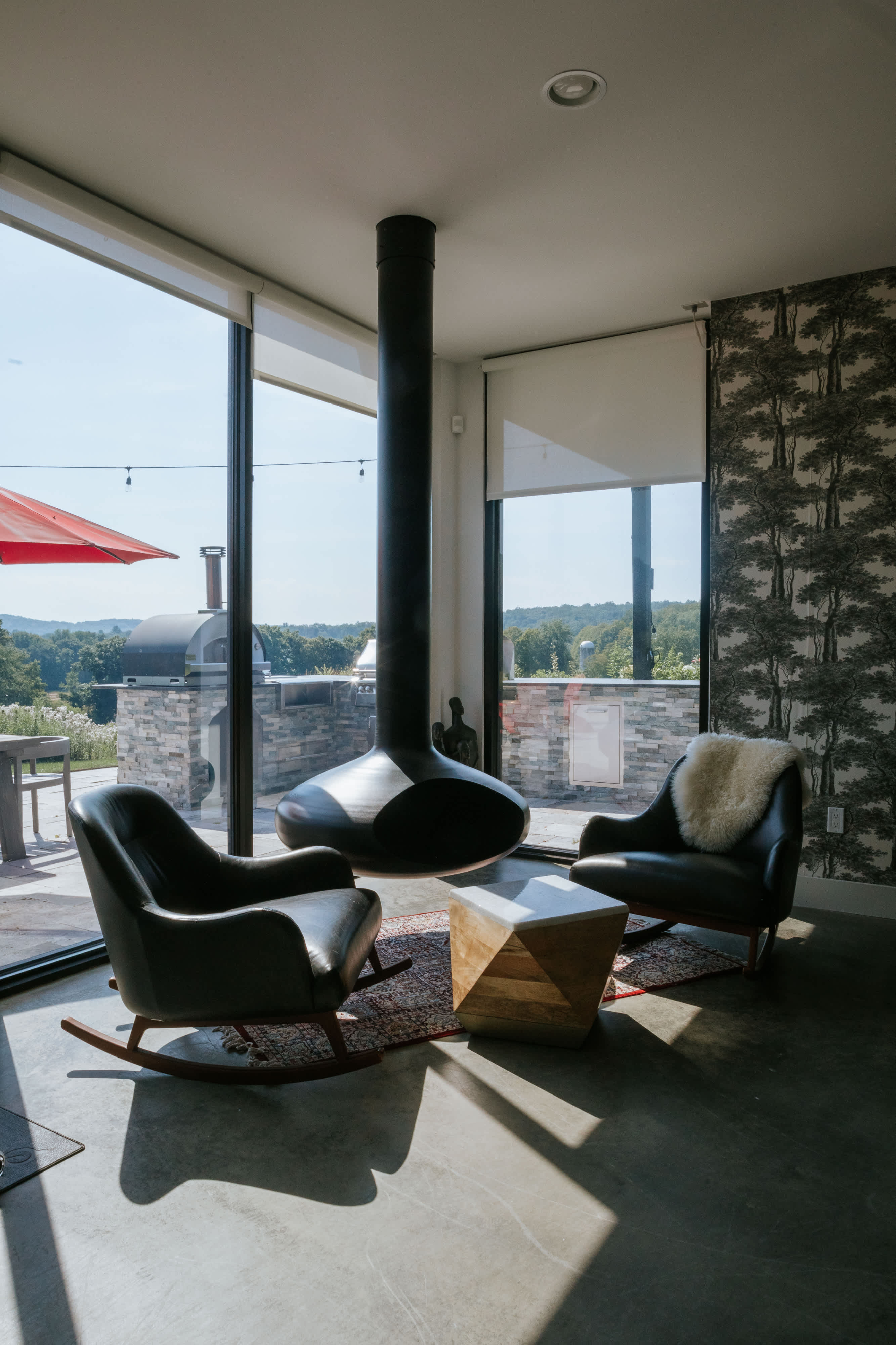 A bright lounge area features two black leather rocking chairs with a wooden geometric side table, centered around a unique black suspended fireplace. Floor-to-ceiling windows offer expansive views of the outdoor landscape and patio.