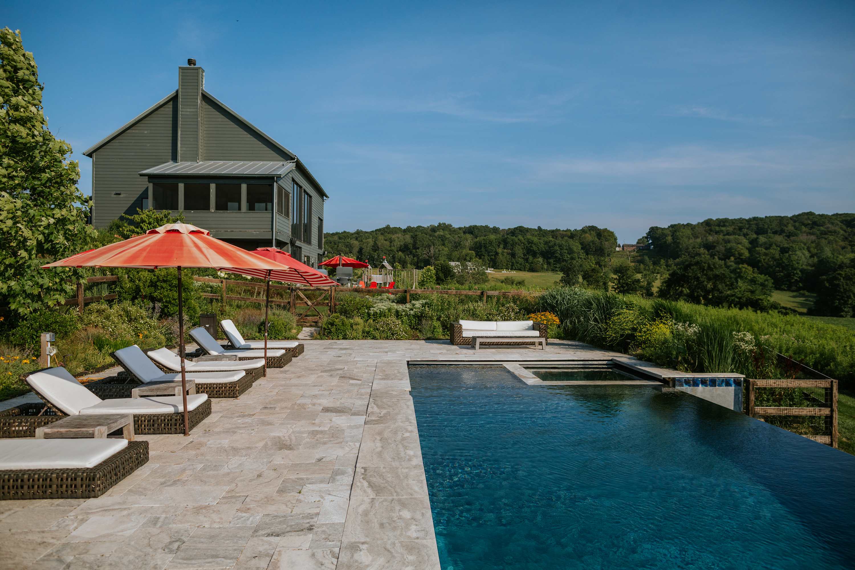 Expansive outdoor pool area featuring an infinity pool, hot tub, multiple lounge chairs with red umbrellas, and a wicker sofa, all set on a stone patio with a large gray house in the background and panoramic views of fields and forests.