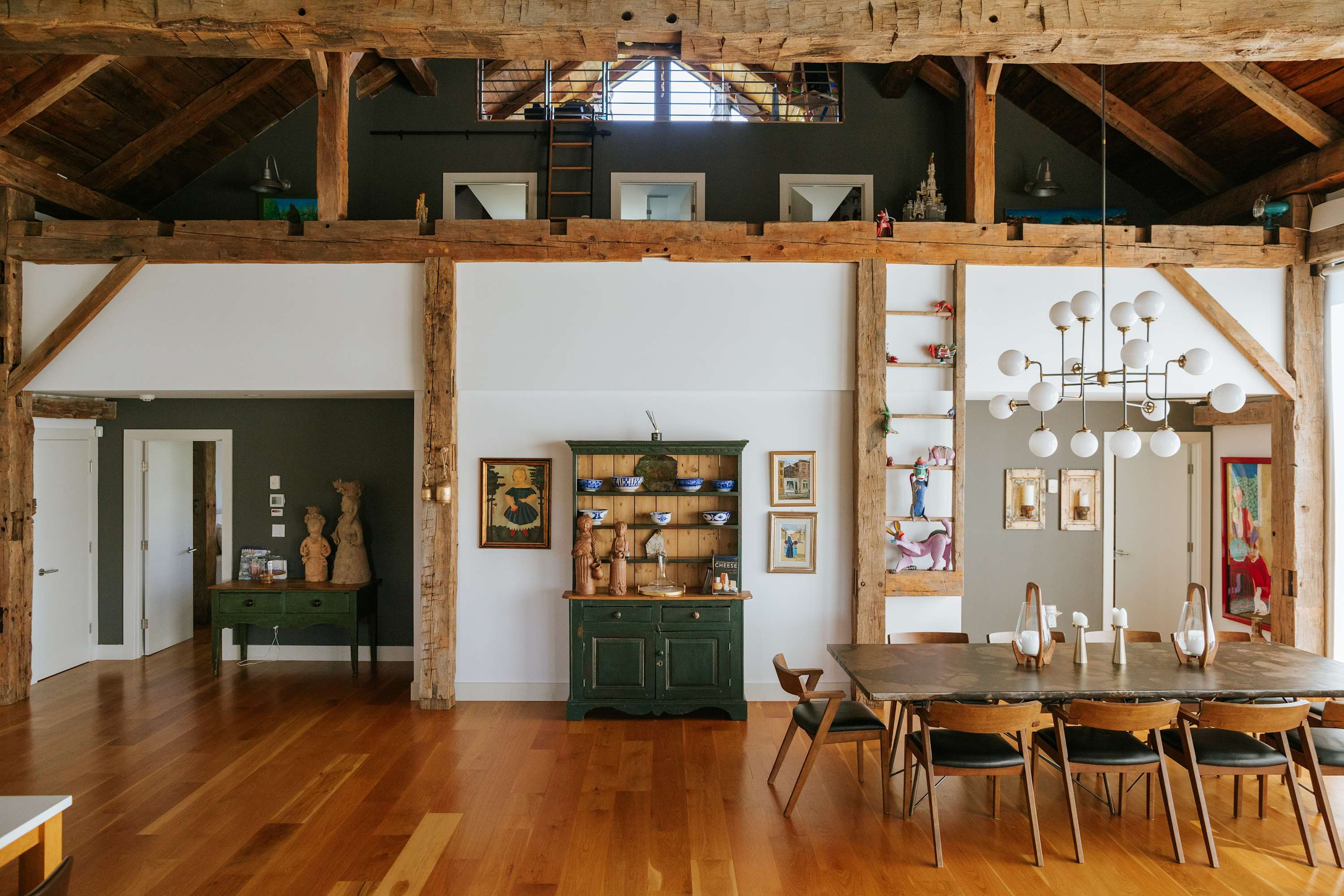 Expansive open-plan dining area featuring a long wooden table with modern chairs, set under a vaulted ceiling with rustic exposed timber beams. A dark green hutch and various artworks adorn the white and dark gray walls, with a loft visible above.