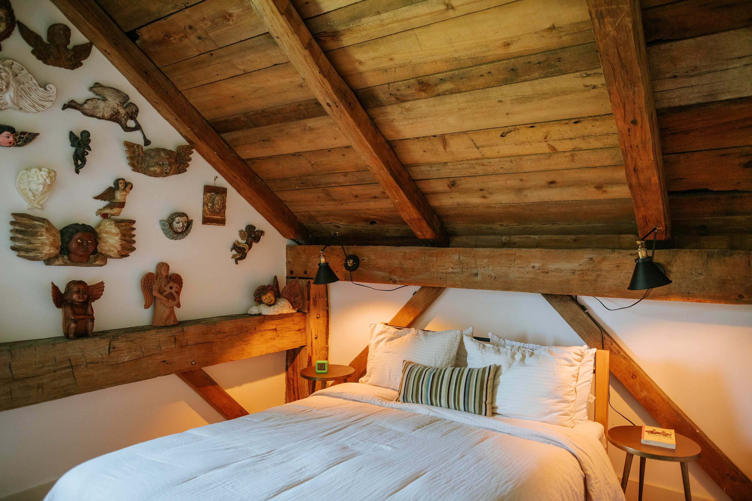 A cozy bedroom showcases a queen bed with white linens and a striped pillow, set against a white wall adorned with various angel-themed sculptures. Exposed wooden beams create a unique headboard and ceiling, with small lamps on wooden nightstands.