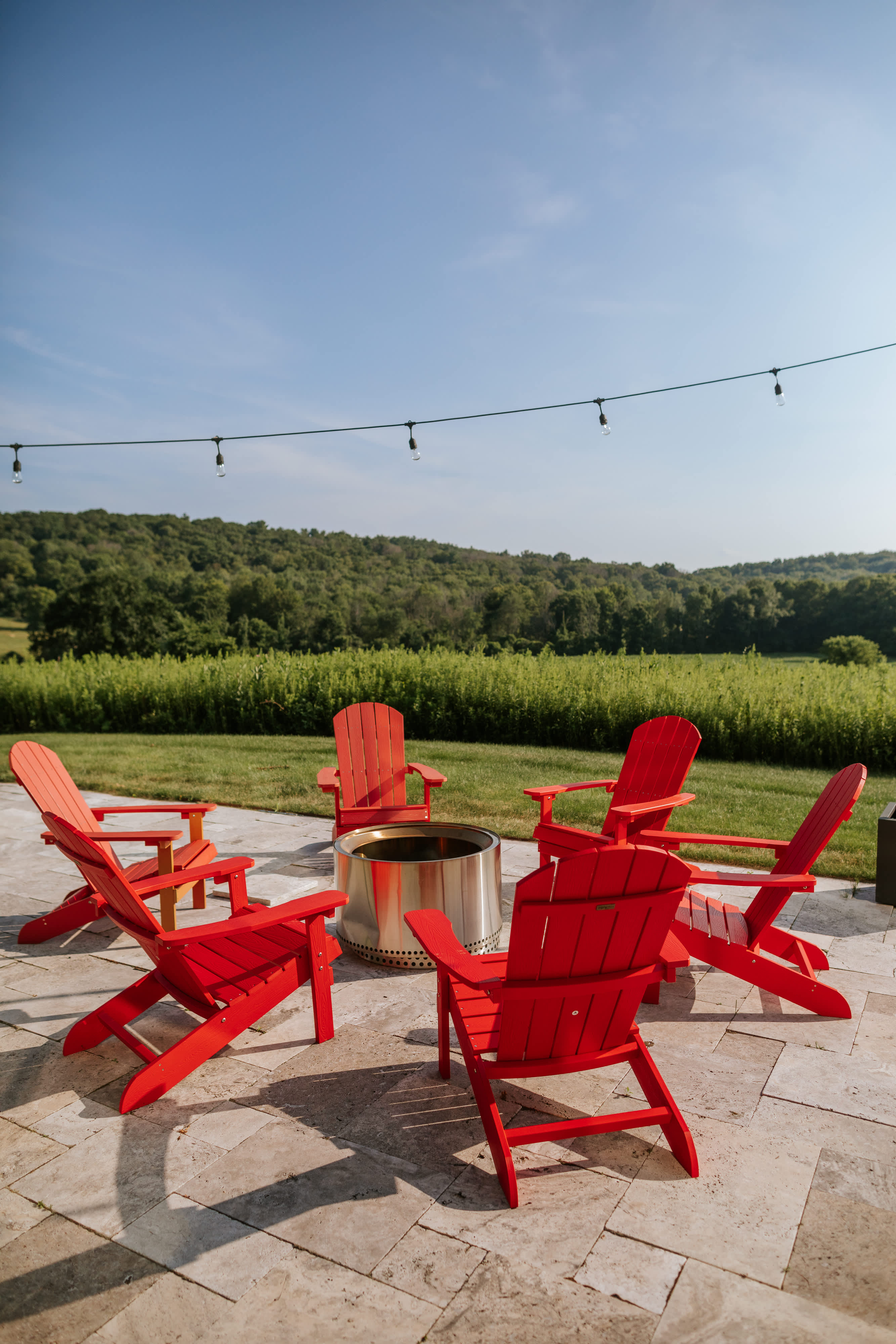 Outdoor fire pit area on a stone patio, featuring a circular arrangement of six bright red Adirondack chairs around a modern, cylindrical metal fire pit, with string lights overhead and green hills in the background.