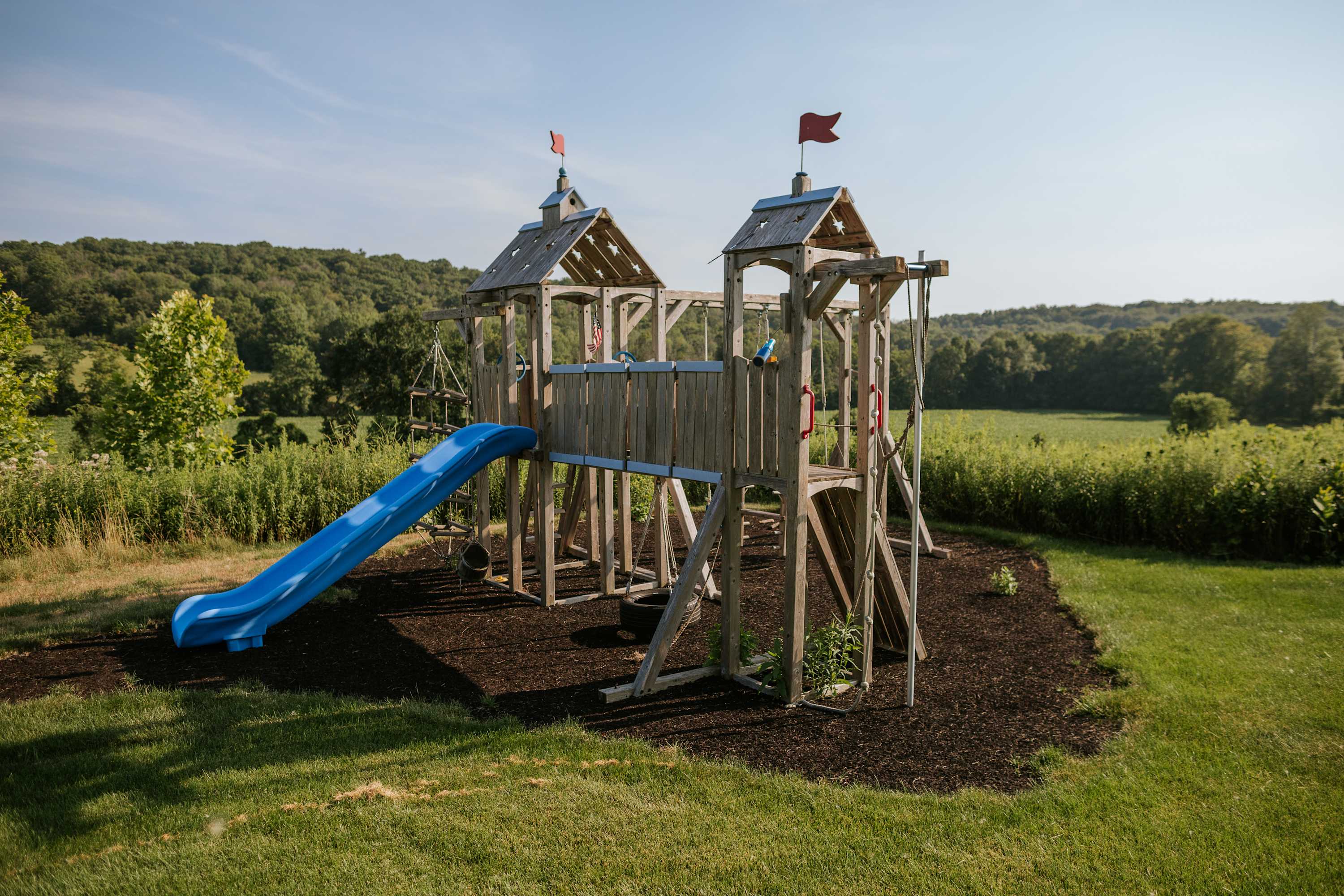 Wooden playground structure with multiple towers, a blue slide, swings, and red flags, set on a bed of dark mulch in a grassy area with a forested background under a clear sky.