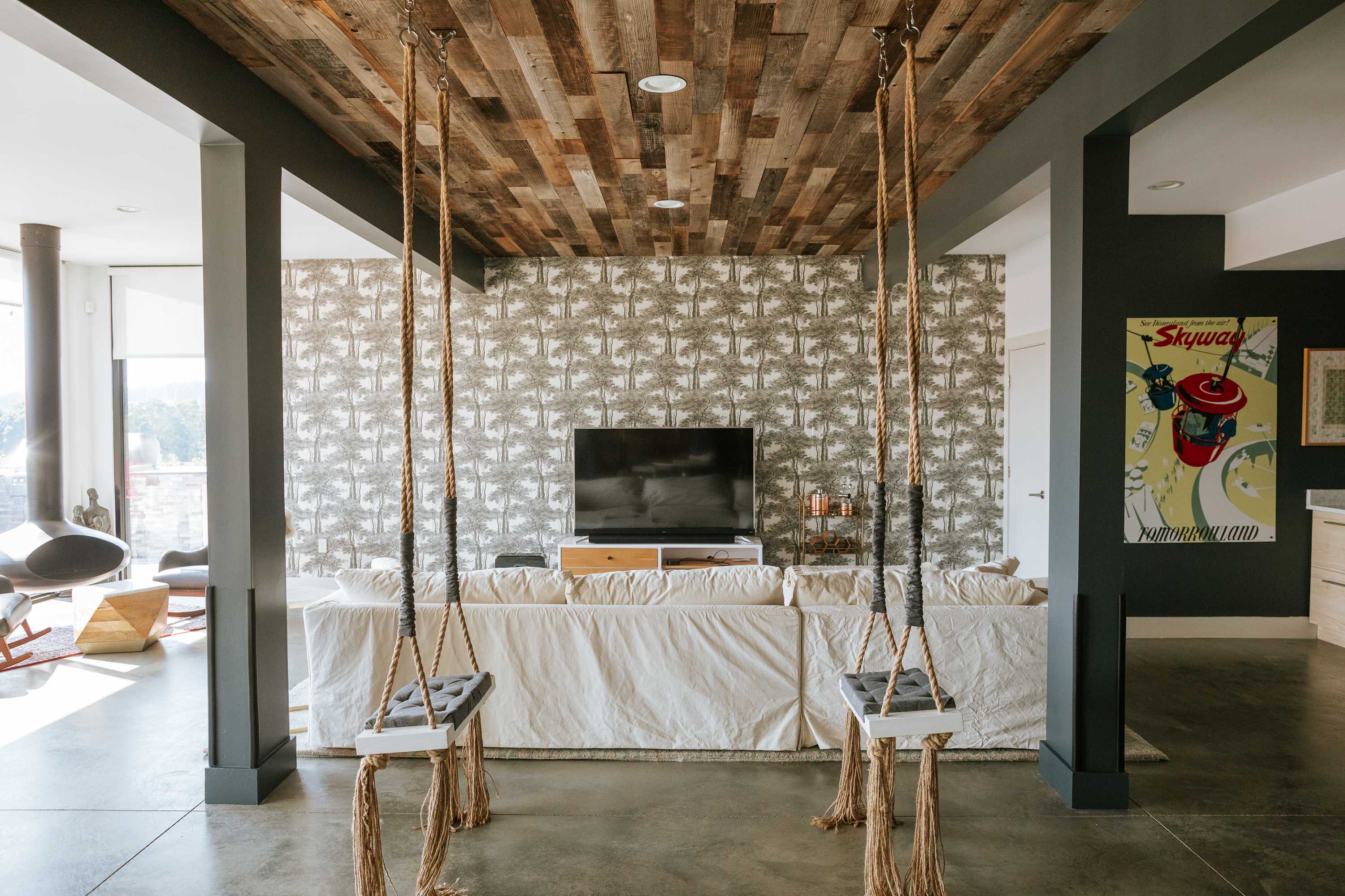 A modern living room features a large white slipcovered sofa facing a TV, set against a palm tree patterned accent wall. Two unique indoor swings hang from the ceiling, which is clad in reclaimed wood, complementing the dark gray structural beams.