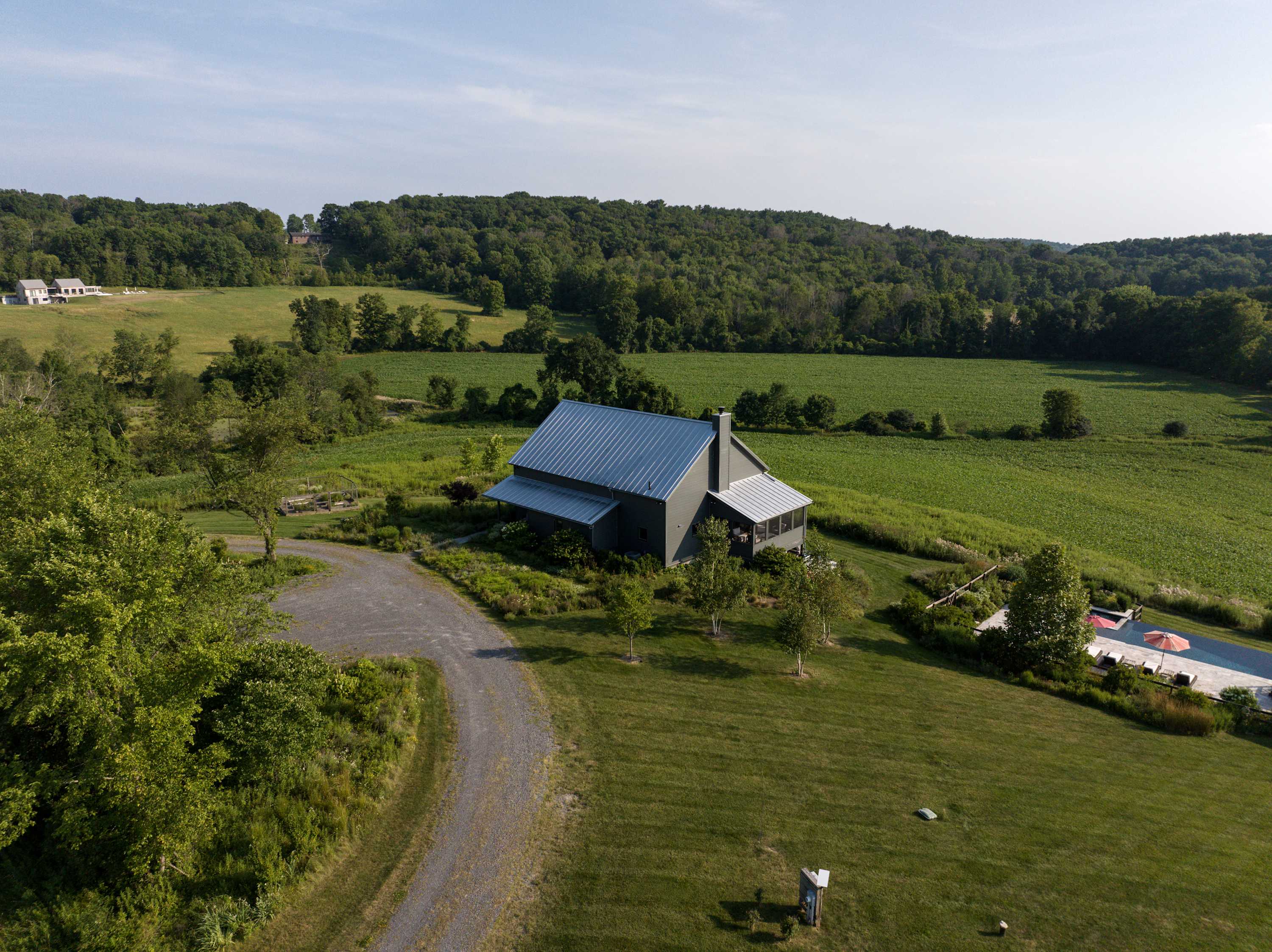 Aerial view of the gray barn-style house with a metal roof, surrounded by expansive green fields and trees, featuring a gravel driveway leading to the property and a glimpse of the pool area to the side.