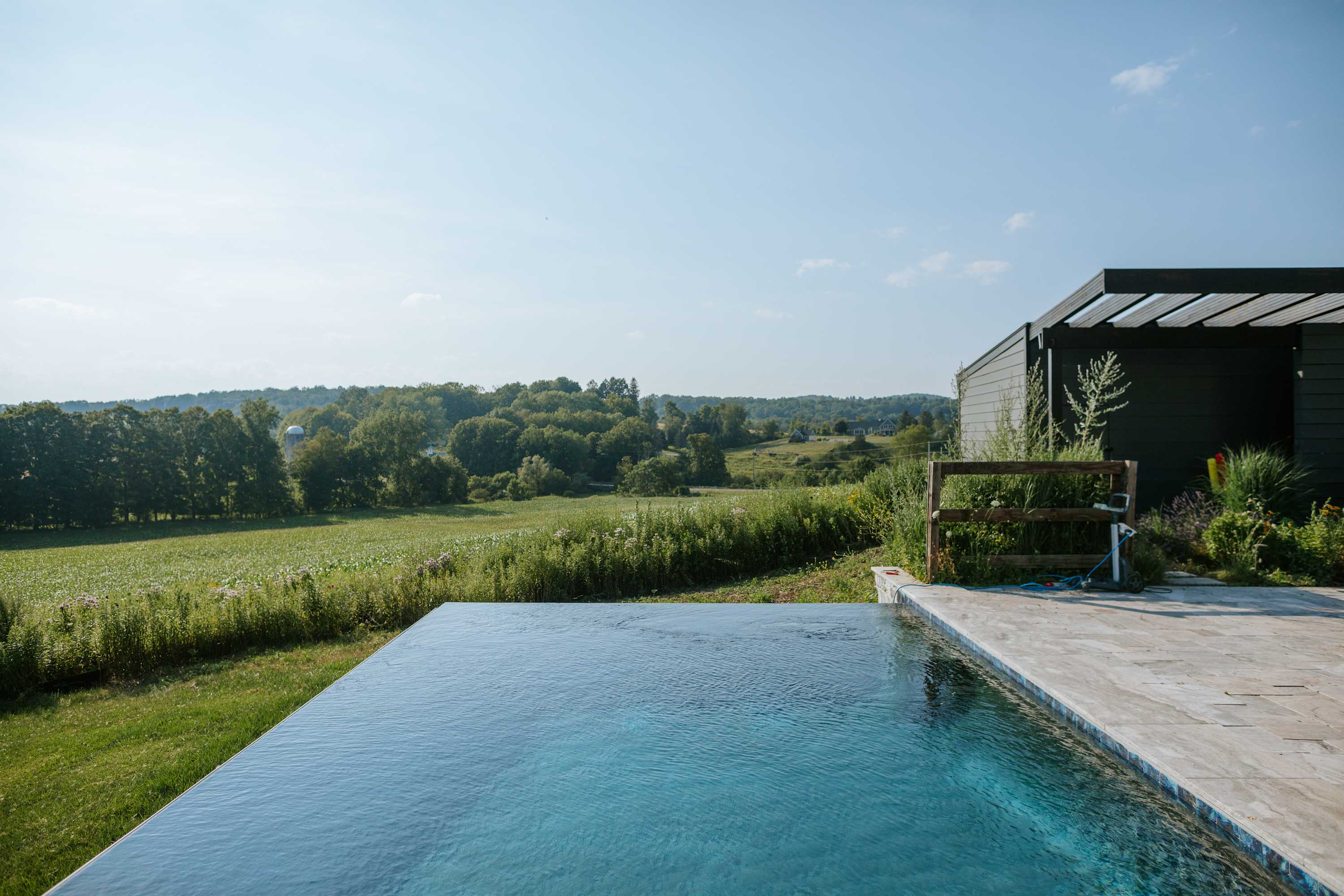 Infinity pool with a view of a modern dark-clad building with a pergola, set against a backdrop of a vast green field, distant trees, and rolling hills under a bright sky.