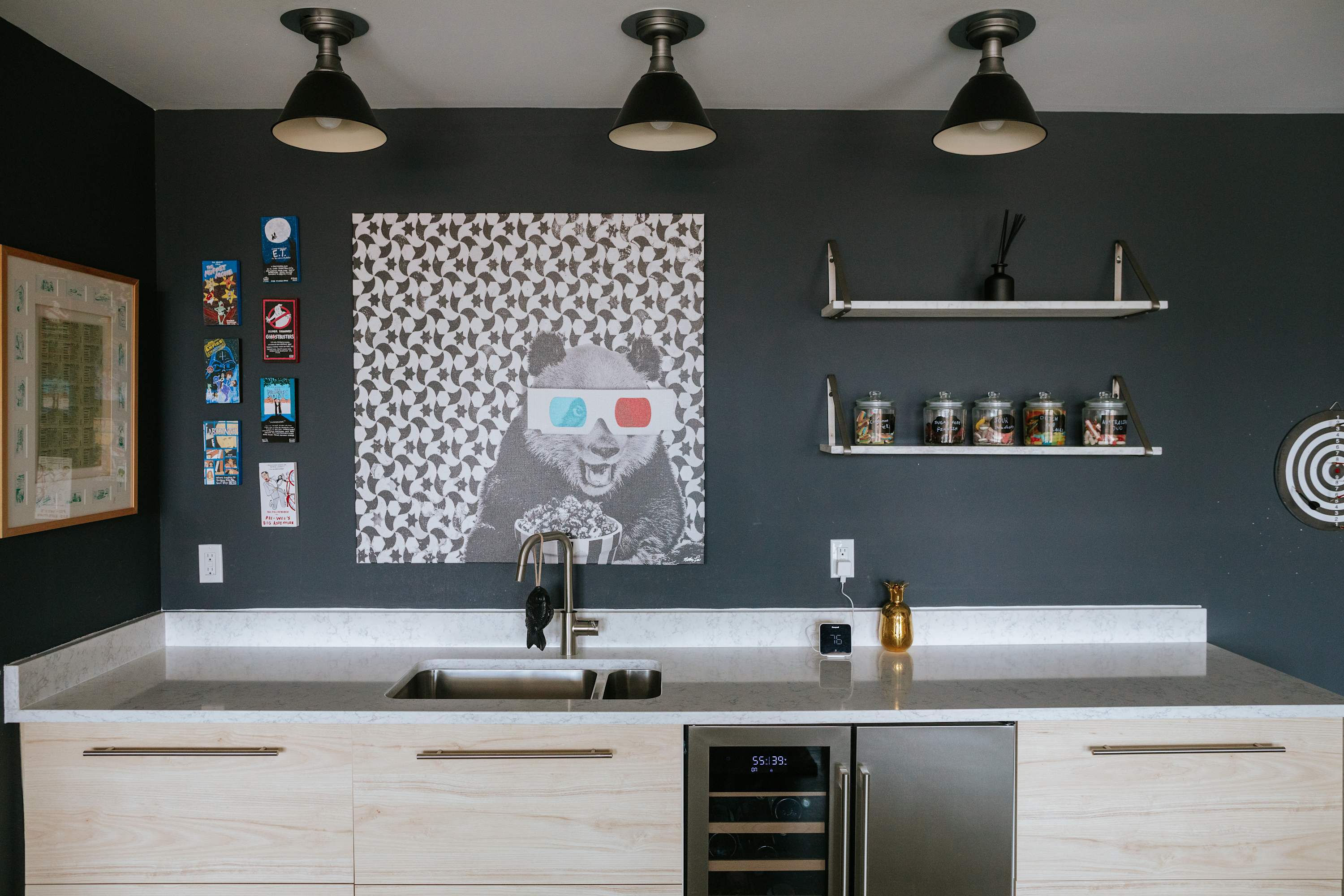 A stylish wet bar area features light wood cabinetry, a white countertop with an undermount sink, and a beverage cooler. The dark gray wall is adorned with a large panda artwork and shelves holding candy jars, illuminated by three industrial pendant lights.