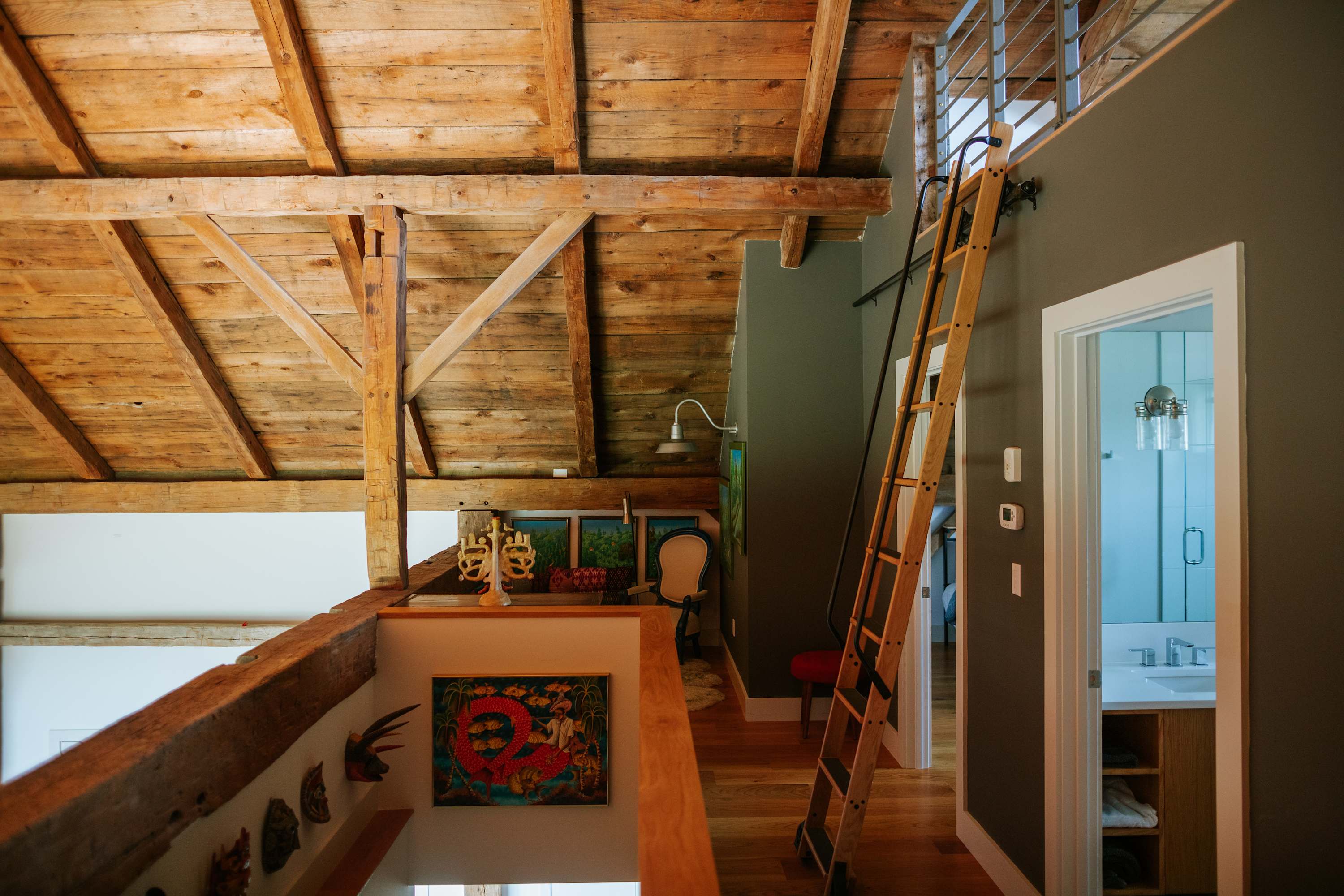 Upper landing or loft area showcasing rustic exposed timber beams and ceiling, with a wooden ladder providing access to an even higher level. Various artworks decorate the walls, and a glimpse of a bathroom is visible through an open doorway.