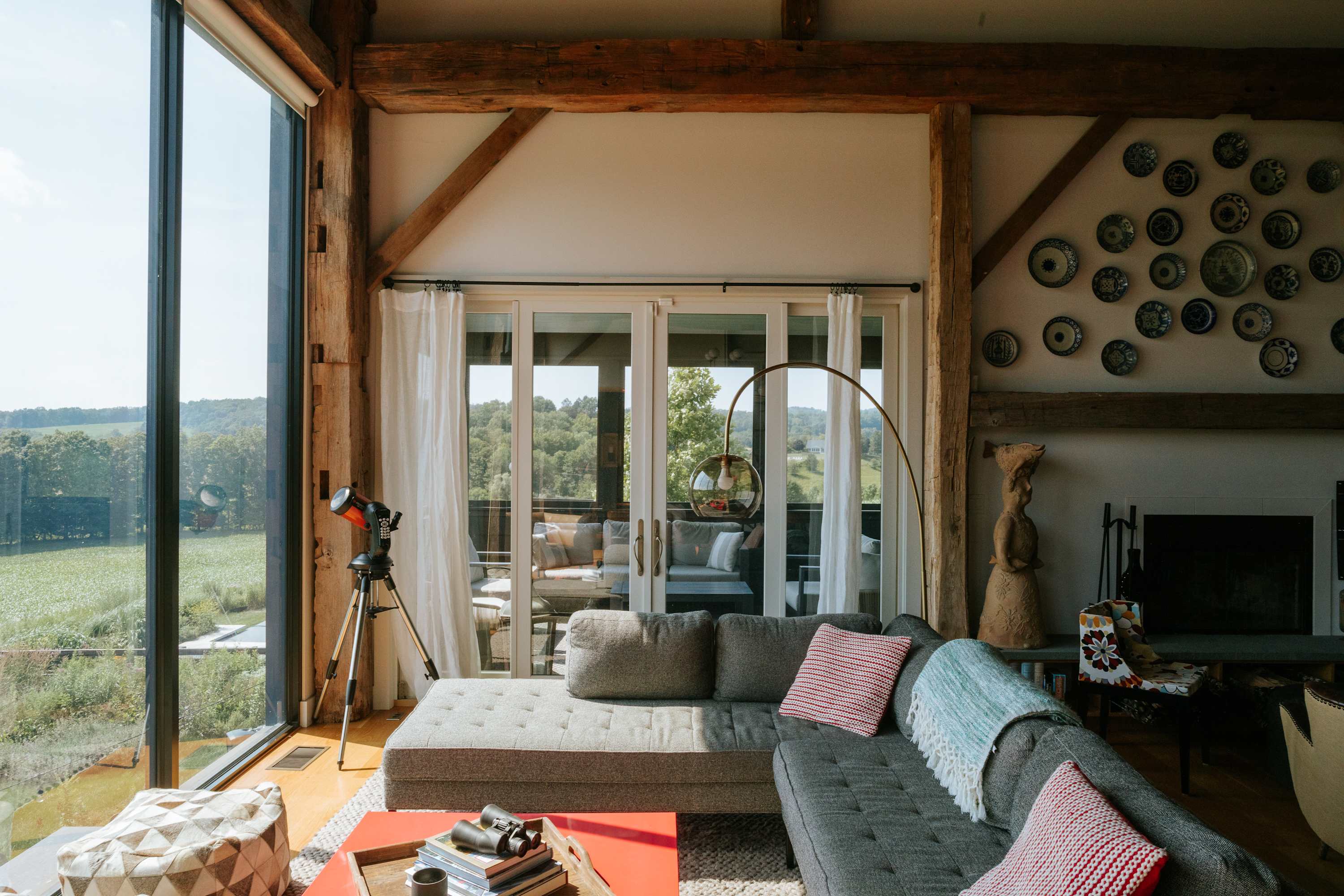 Bright living room with a large gray sectional sofa and a red coffee table, offering panoramic views of rolling hills through expansive floor-to-ceiling windows. White curtains frame glass doors leading to an outdoor patio, and a telescope stands ready for stargazing.