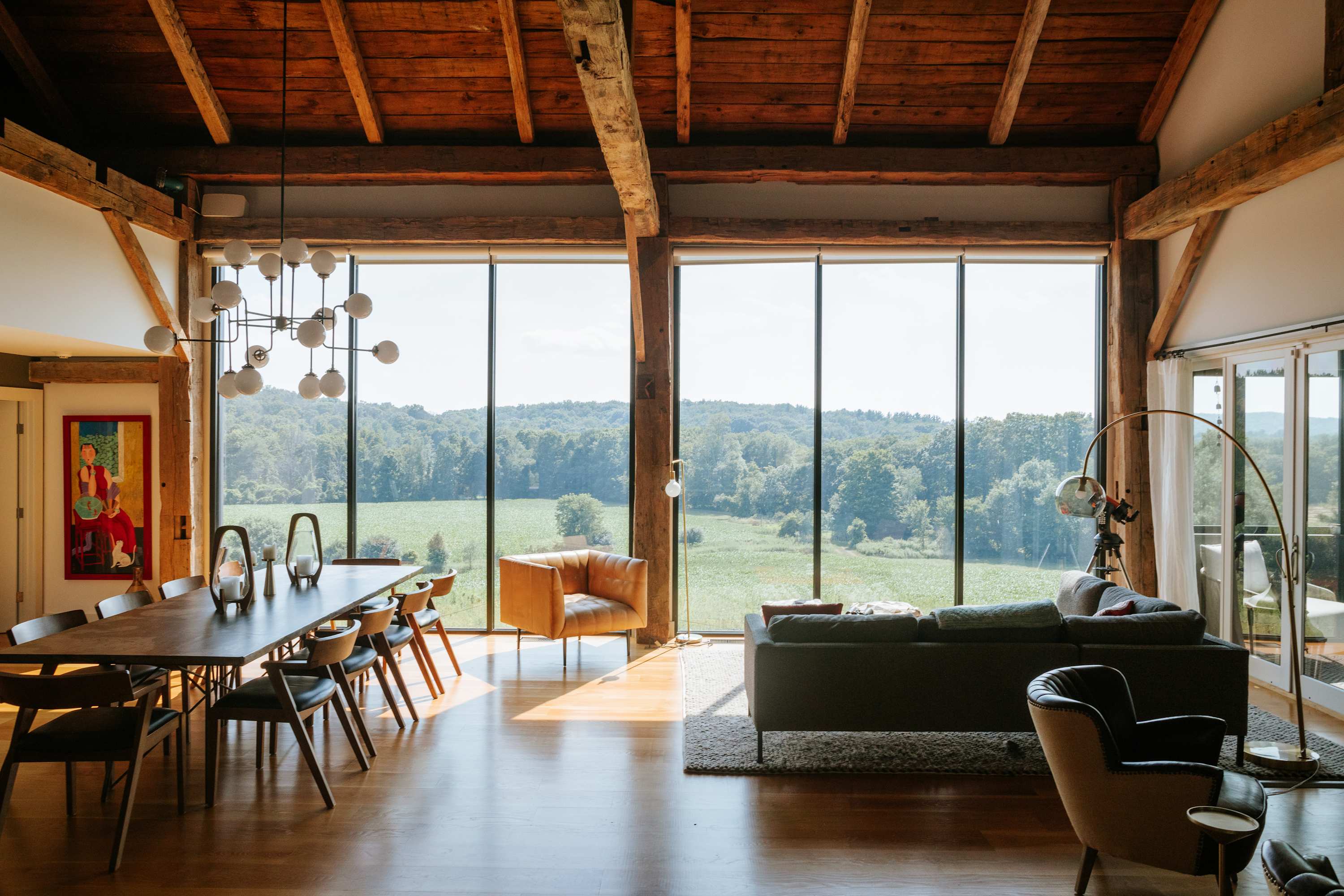 A spacious living and dining area boasts floor-to-ceiling windows offering panoramic views of the surrounding green landscape, featuring a long wooden dining table with modern chairs, a large sofa, and exposed wooden beams overhead.