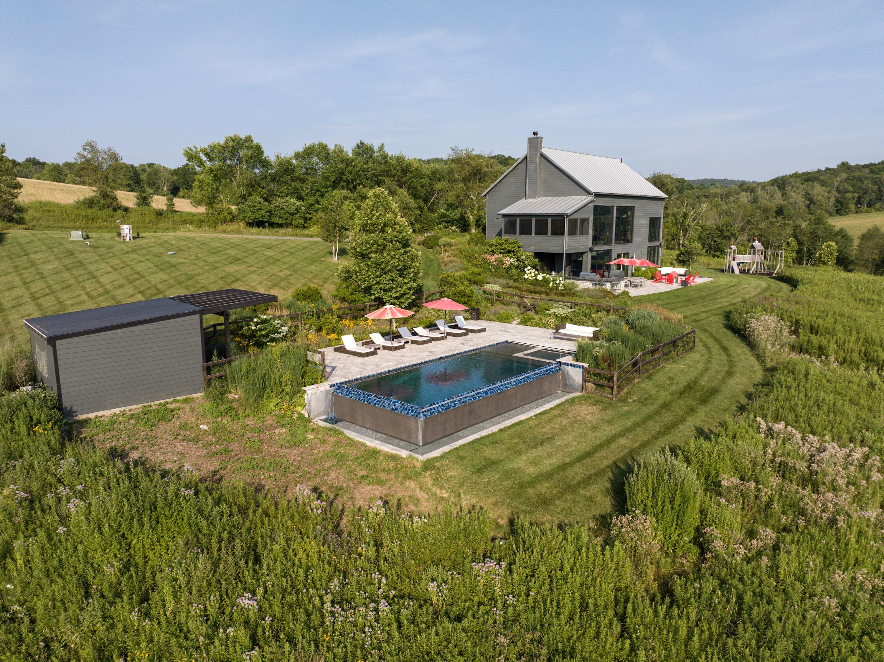 Aerial view capturing the modern gray house, the infinity-edge swimming pool with its surrounding patio and lounge chairs, and a wooden playground, all situated on a grassy hill overlooking expansive green fields.
