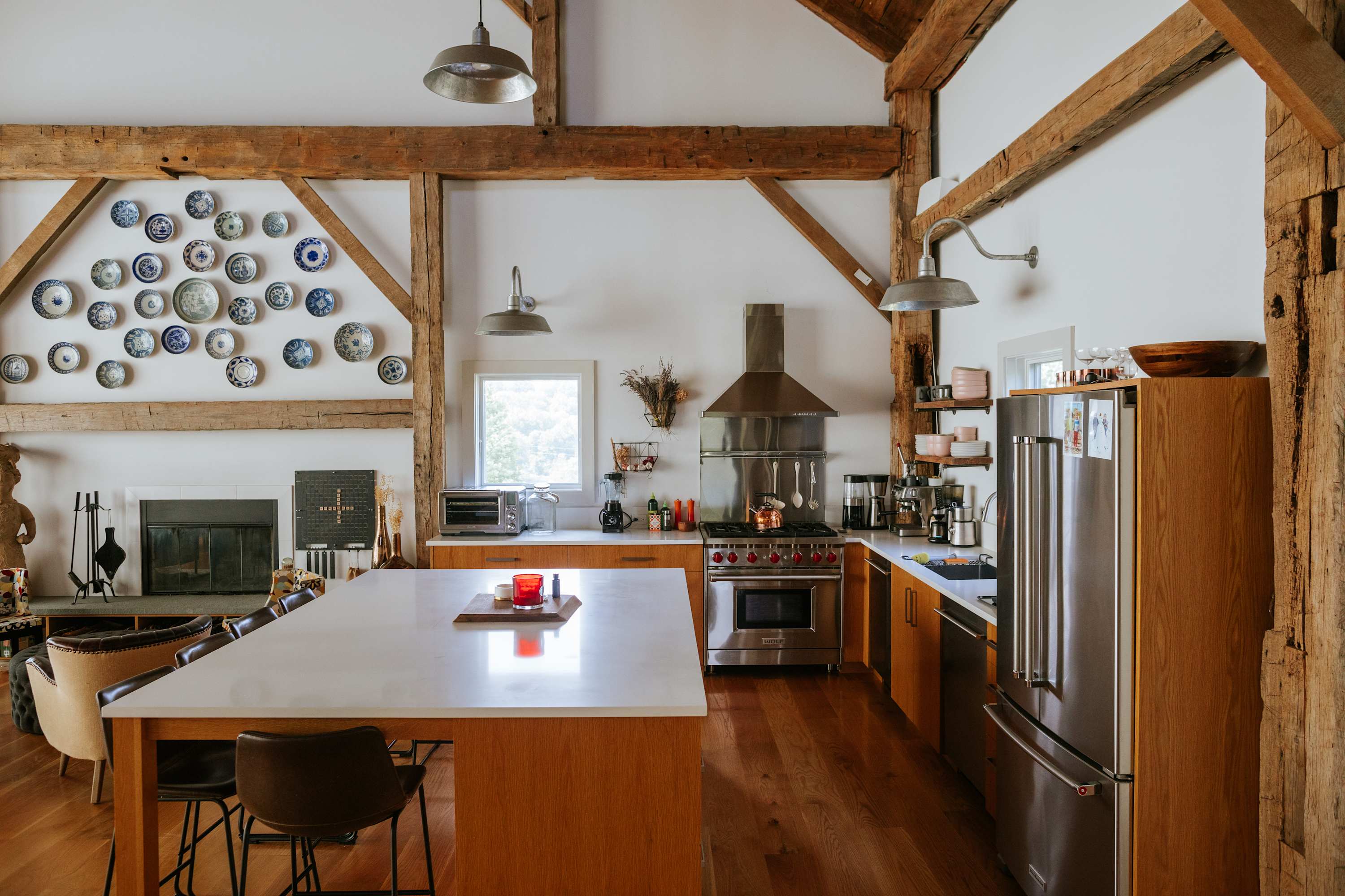 A well-appointed kitchen features light wood cabinetry, a large white island with bar stools, and professional-grade stainless steel appliances, all integrated into an open-concept space with exposed wooden beams and white walls.