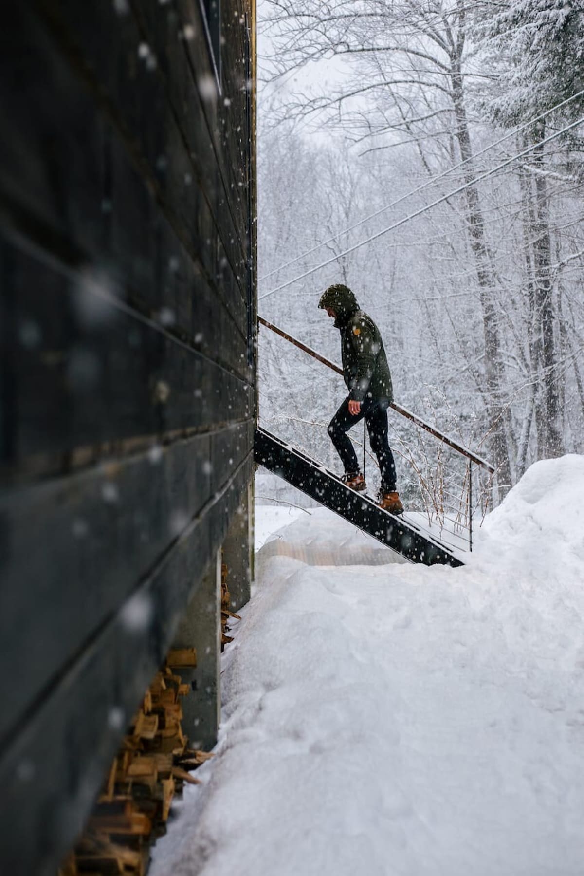 A person bundled in winter gear ascends the exterior metal stairs of the cabin during a heavy snowfall, with deep snowdrifts covering the ground. Stacked firewood is visible along the dark wood-sided exterior of the house.