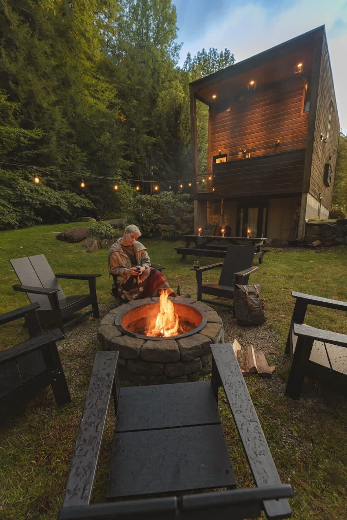 Outdoor fire pit area with a stone fire pit surrounded by black Adirondack chairs, string lights, and lush green trees, with the modern cabin visible in the background.