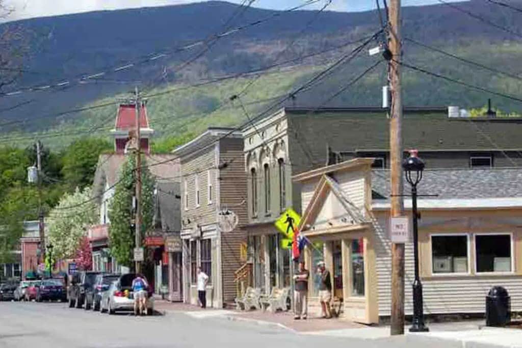 Quaint small-town street lined with historic buildings housing shops, with people walking on the sidewalks and cars parked along the curb, all set against a backdrop of lush green mountains.