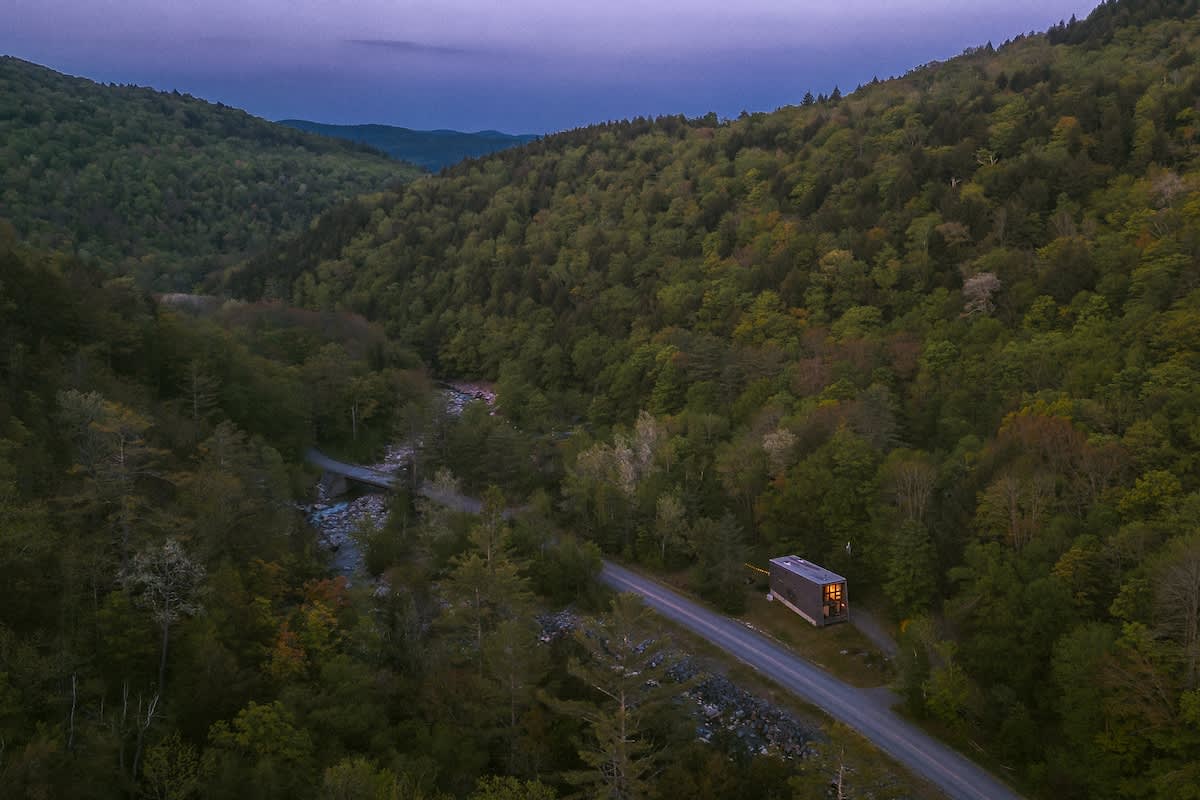 Panoramic aerial view of the modern cabin nestled in a lush green valley beside a winding road and a river, surrounded by dense forest under a twilight sky.