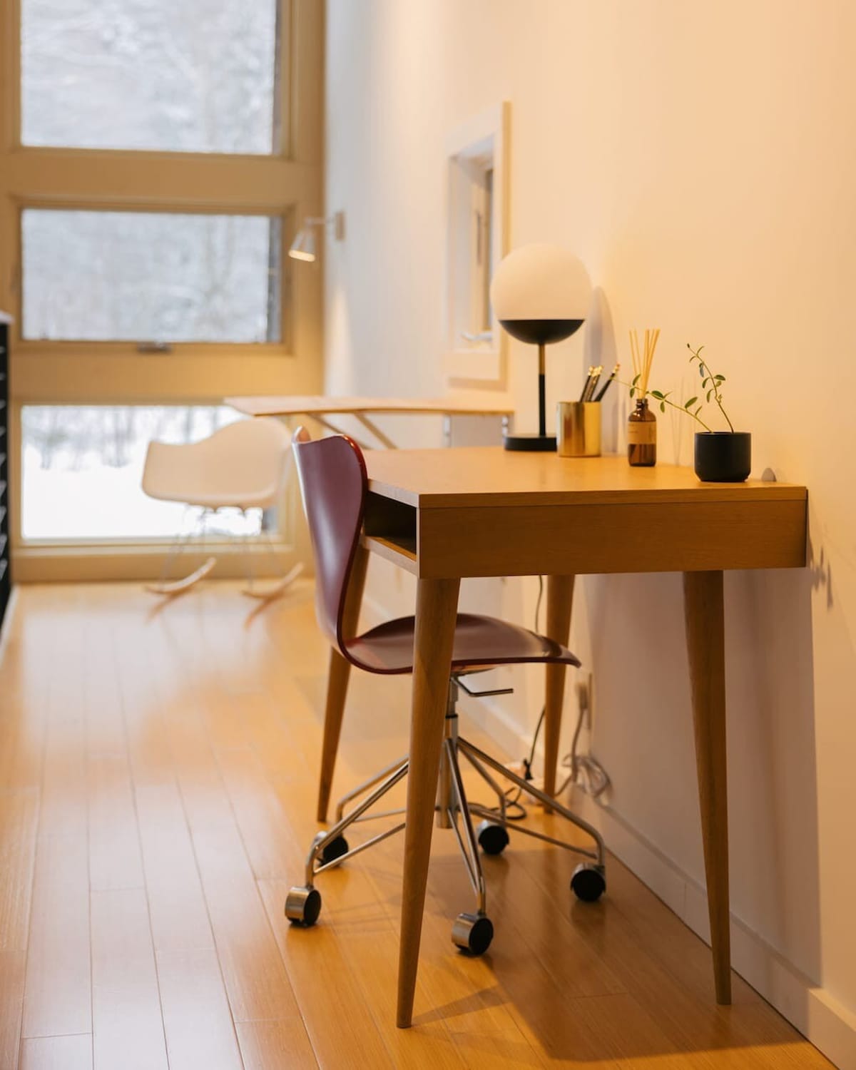 A close-up of a modern wooden desk with tapered legs, a red rolling chair, and a minimalist globe lamp. The desk is adorned with a small plant, pens, and a diffuser, set against a white wall with large windows revealing a snowy outdoor scene.
