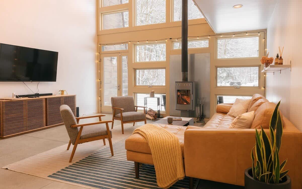 Spacious living room featuring a modern wood-burning stove, a large tan leather sectional sofa, and two grey armchairs on a striped rug. A wall-mounted TV is visible above a slatted wooden media console, and large windows overlook a snowy outdoor scene.